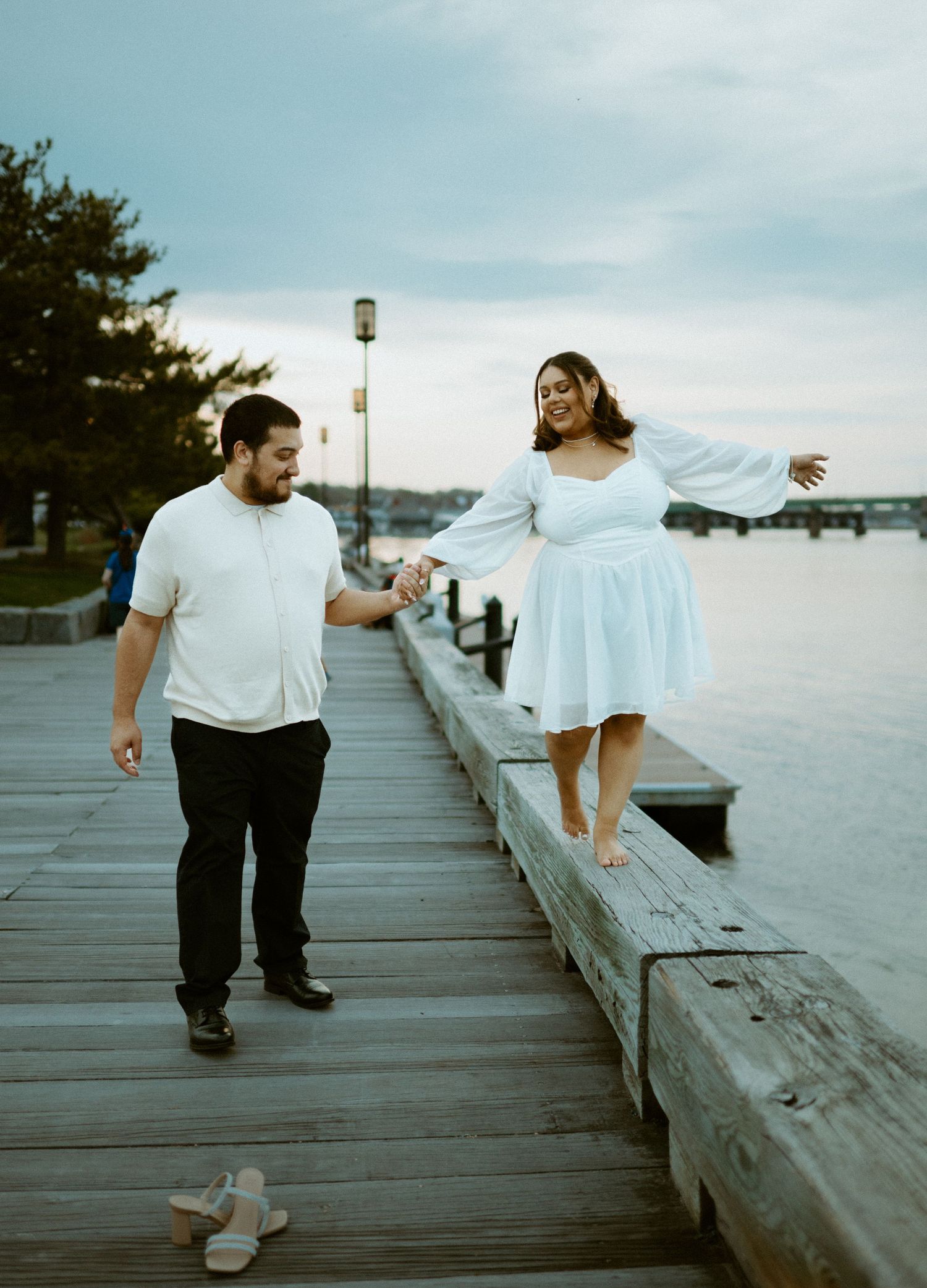 Couple holding hands on a wooden pier at dusk, woman in white dress balancing on the railing by the water.