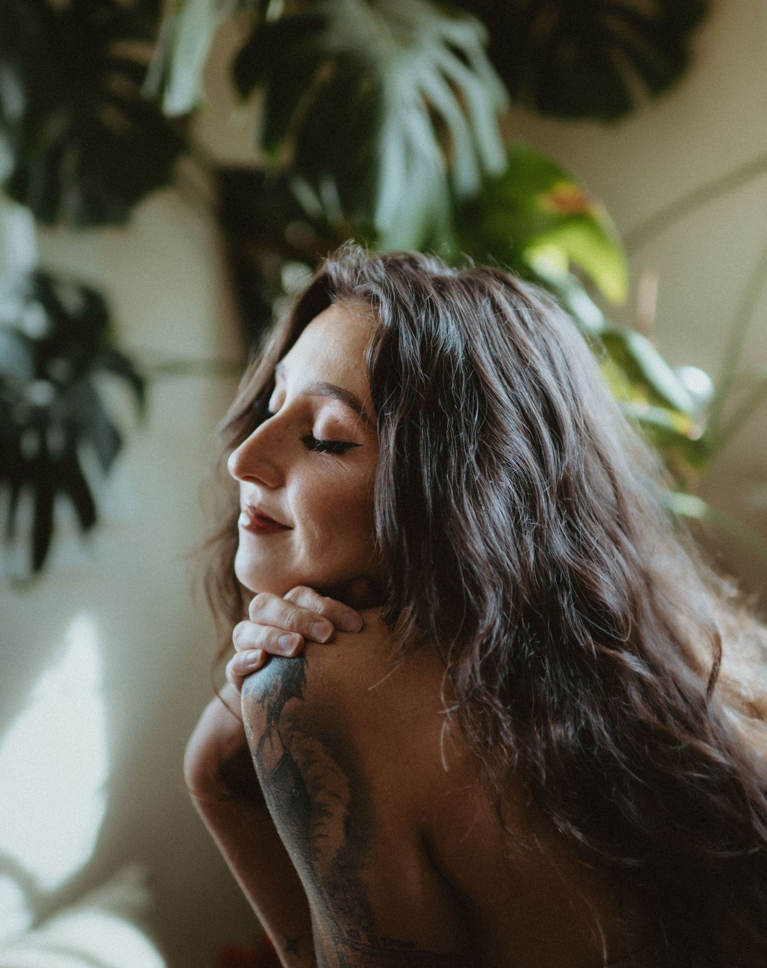 A tattooed woman with long hair poses with eyes closed among lush green monstera plants in soft natural light.