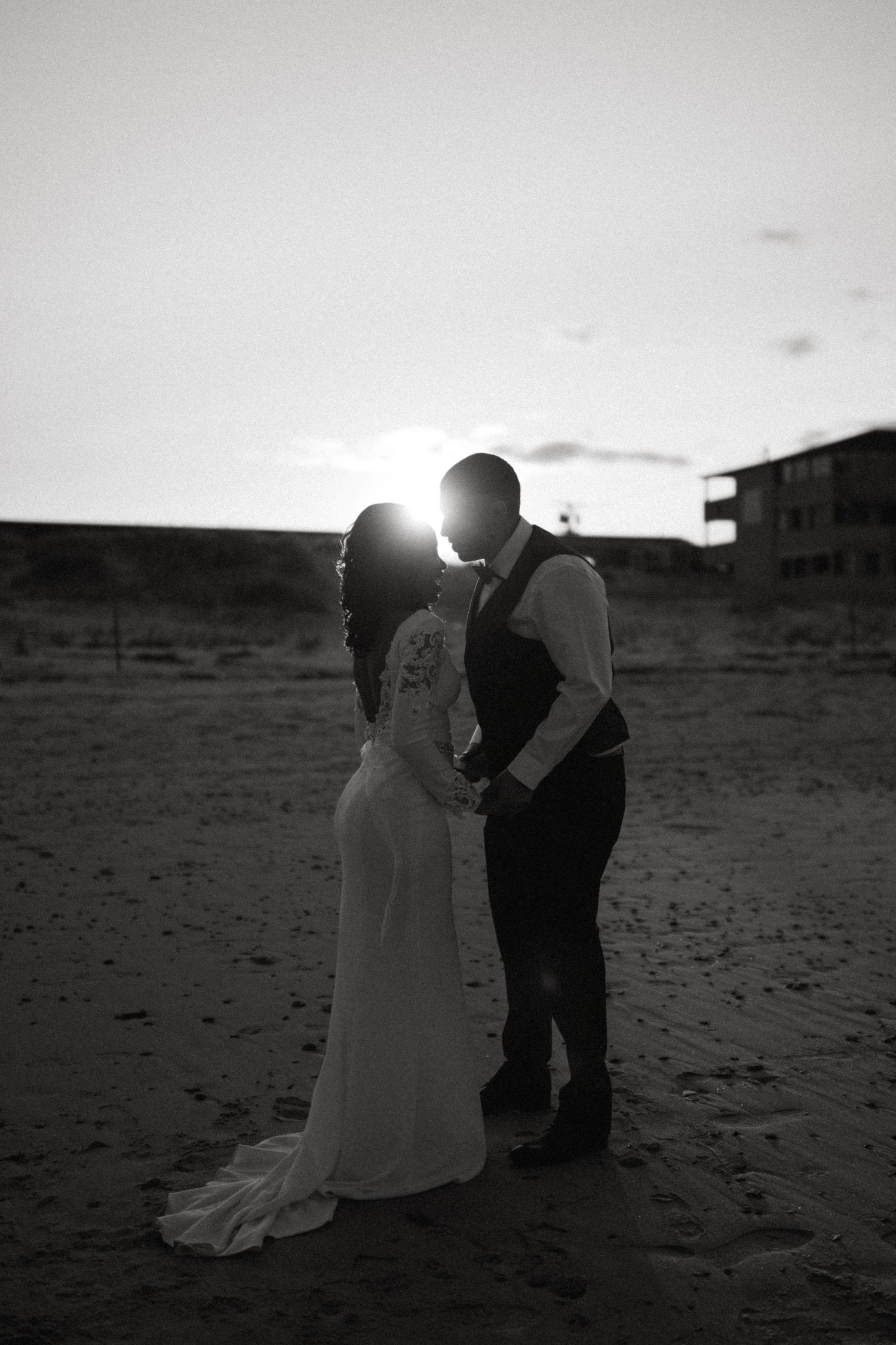 Black and white romantic photo of bride and groom kissing at sunset on sandy beach.