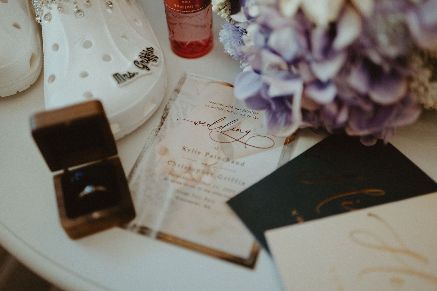 Elegant wedding stationery with gold calligraphy, ring box, and purple hydrangeas on white table.