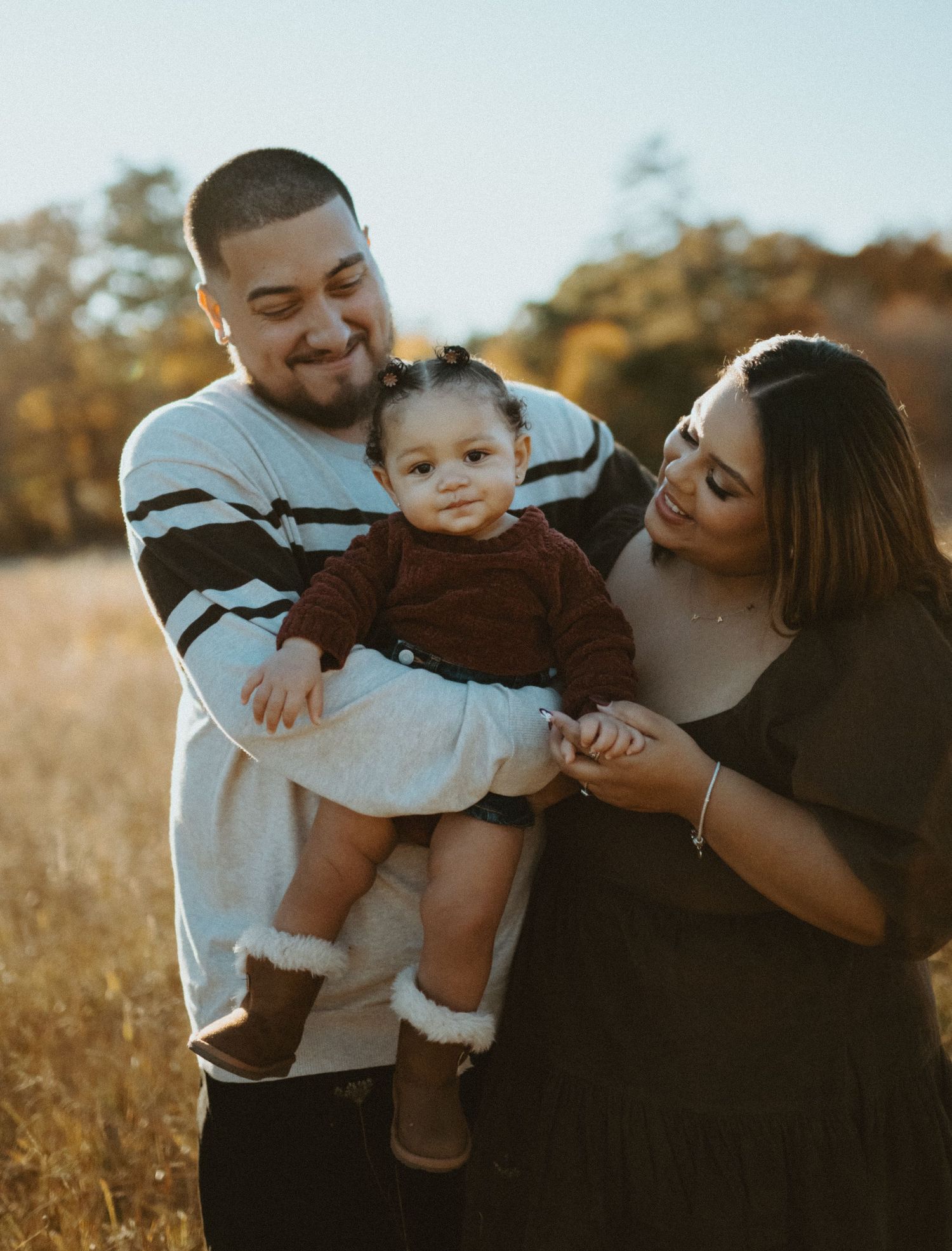 Happy young family holding toddler girl in autumn field during golden hour.