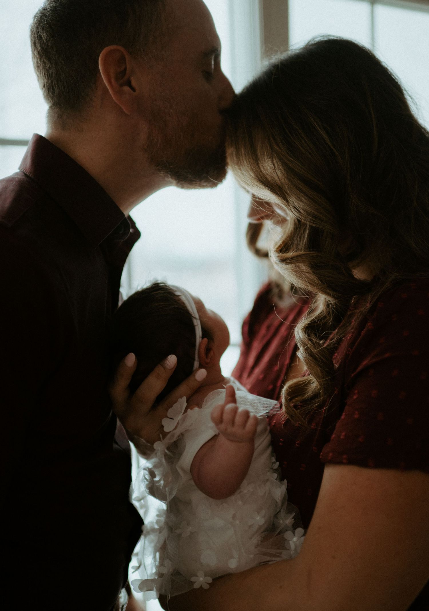 Father kisses mother's forehead while tenderly holding their newborn baby in a white lace gown.