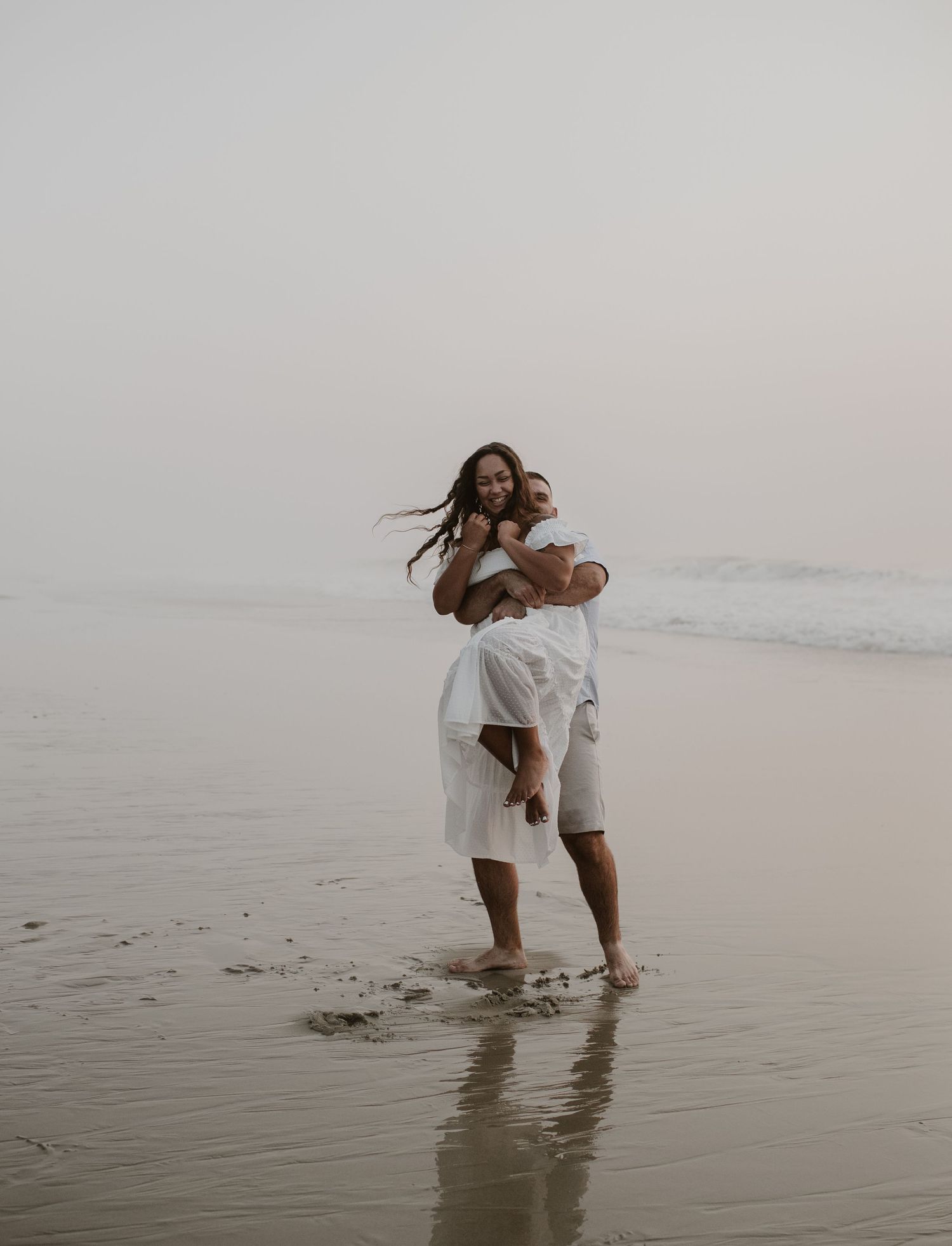 Happy couple embracing on a misty beach, both wearing white outfits, standing in shallow water.