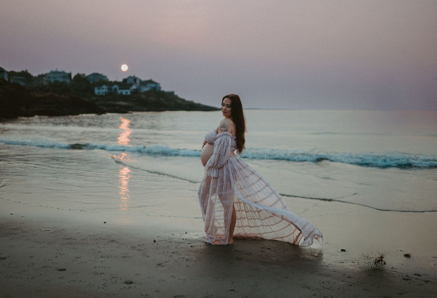 Pregnant woman in pink flowy dress walking on a serene beach during sunrise. Early morning sun looms in the background