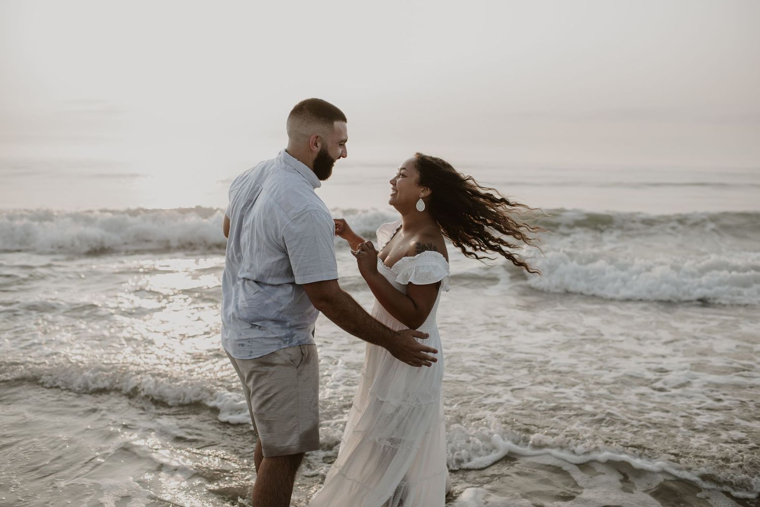 Joyful couple dancing together in ocean waves on the beach, womans hair blowing in the wind.