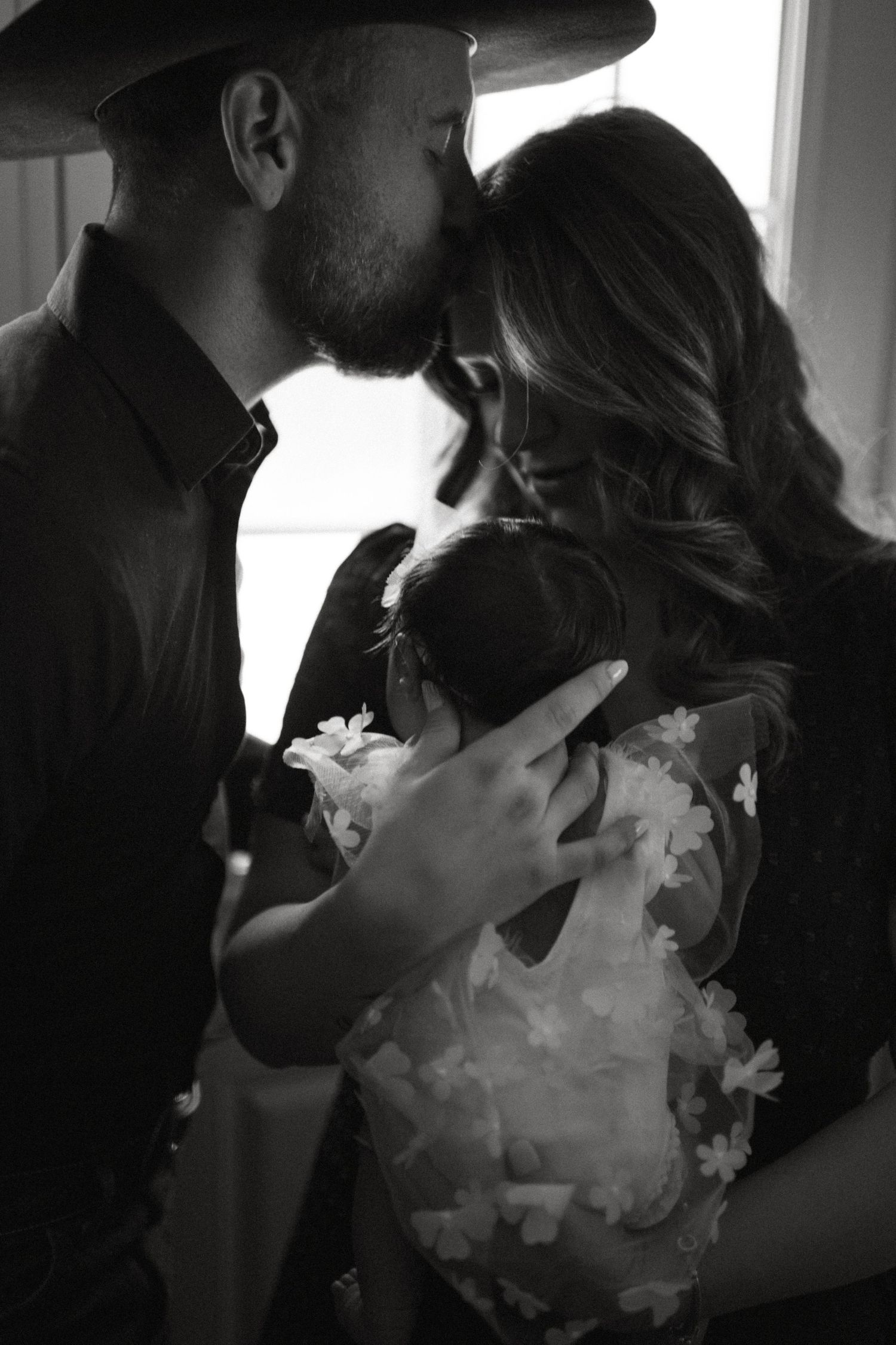 Black and white photo of parents sharing a tender moment while holding their newborn baby together.