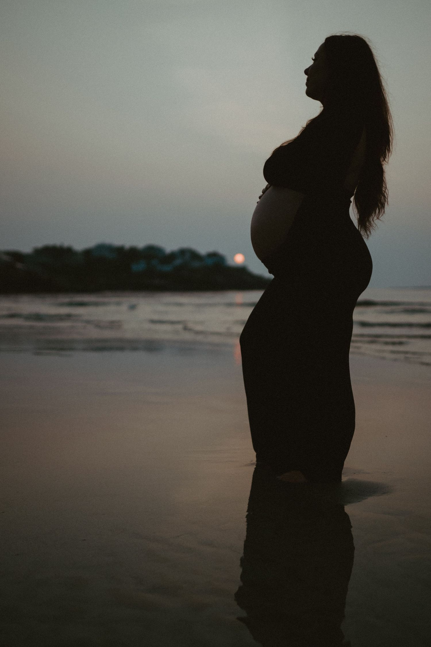 Silhouette of a pregnant woman cradling her belly on a beach at sunrise with calm ocean waves.