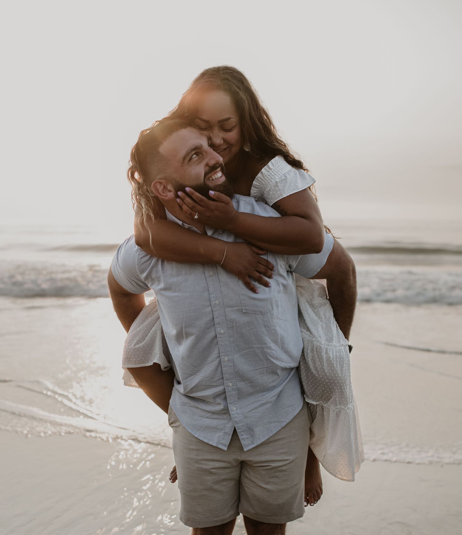 Man giving woman a piggyback ride on the beach, both laughing joyfully near the ocean.