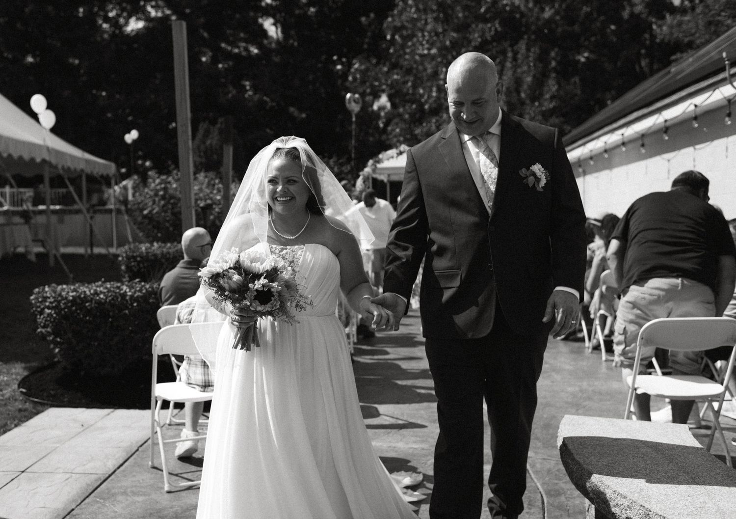Bride and groom walking down aisle at outdoor wedding ceremony in black and white photo.