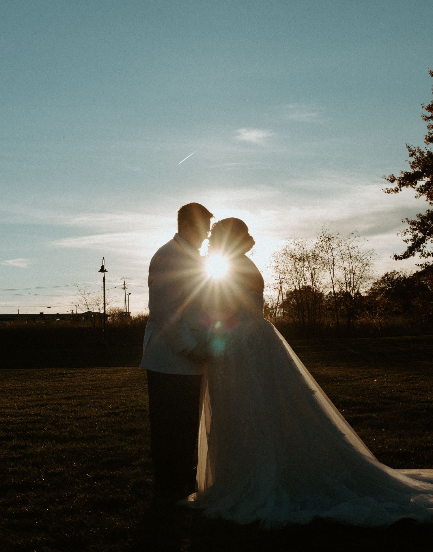 Romantic silhouette of brides with sun flare between them against blue sky at sunset.