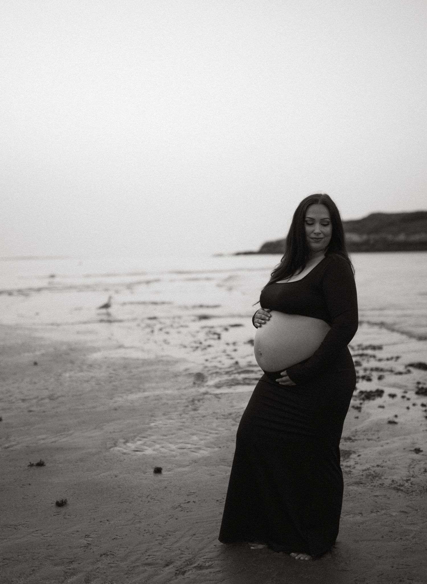 Black and white photo of pregnant woman in black dress cradling baby bump on serene beach.