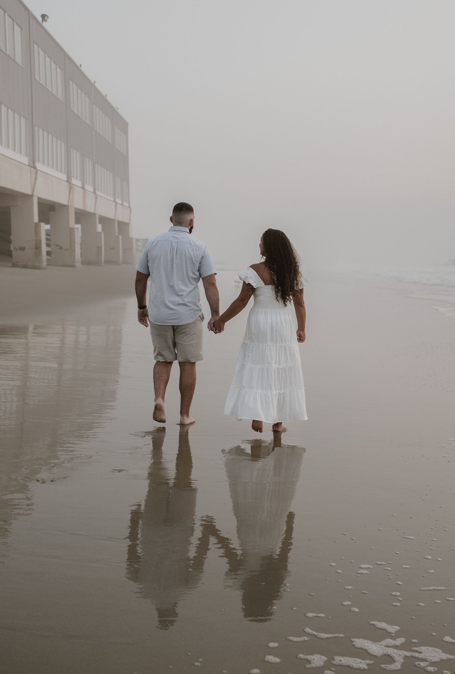 Couple walking hand in hand on reflective wet beach with coastal building in misty background.