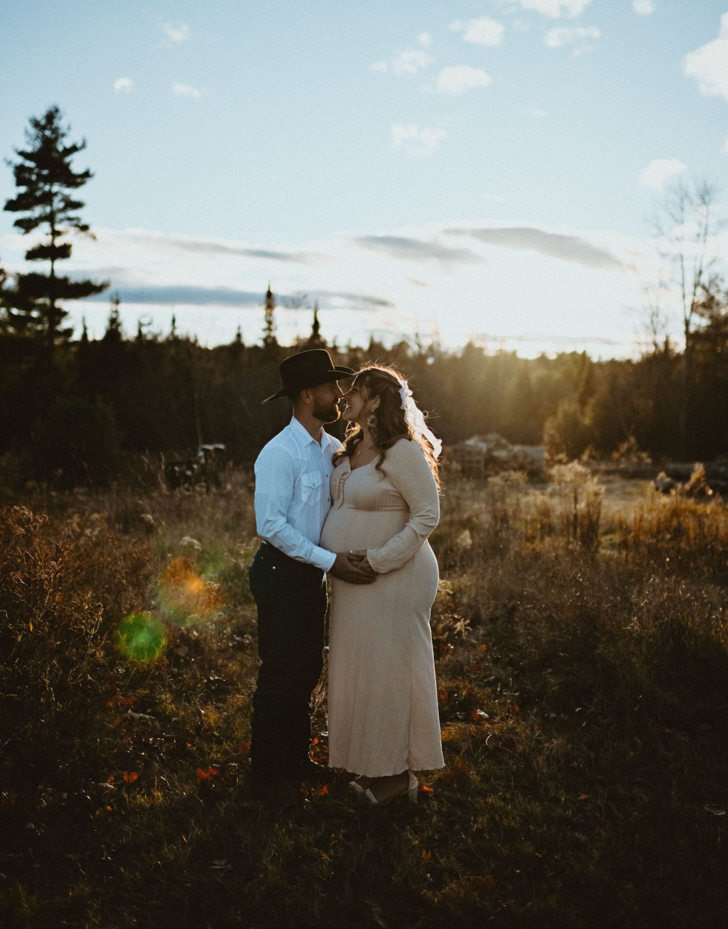 Couple embracing at golden hour during maternity shoot in autumn field, woman in beige maternity dress.