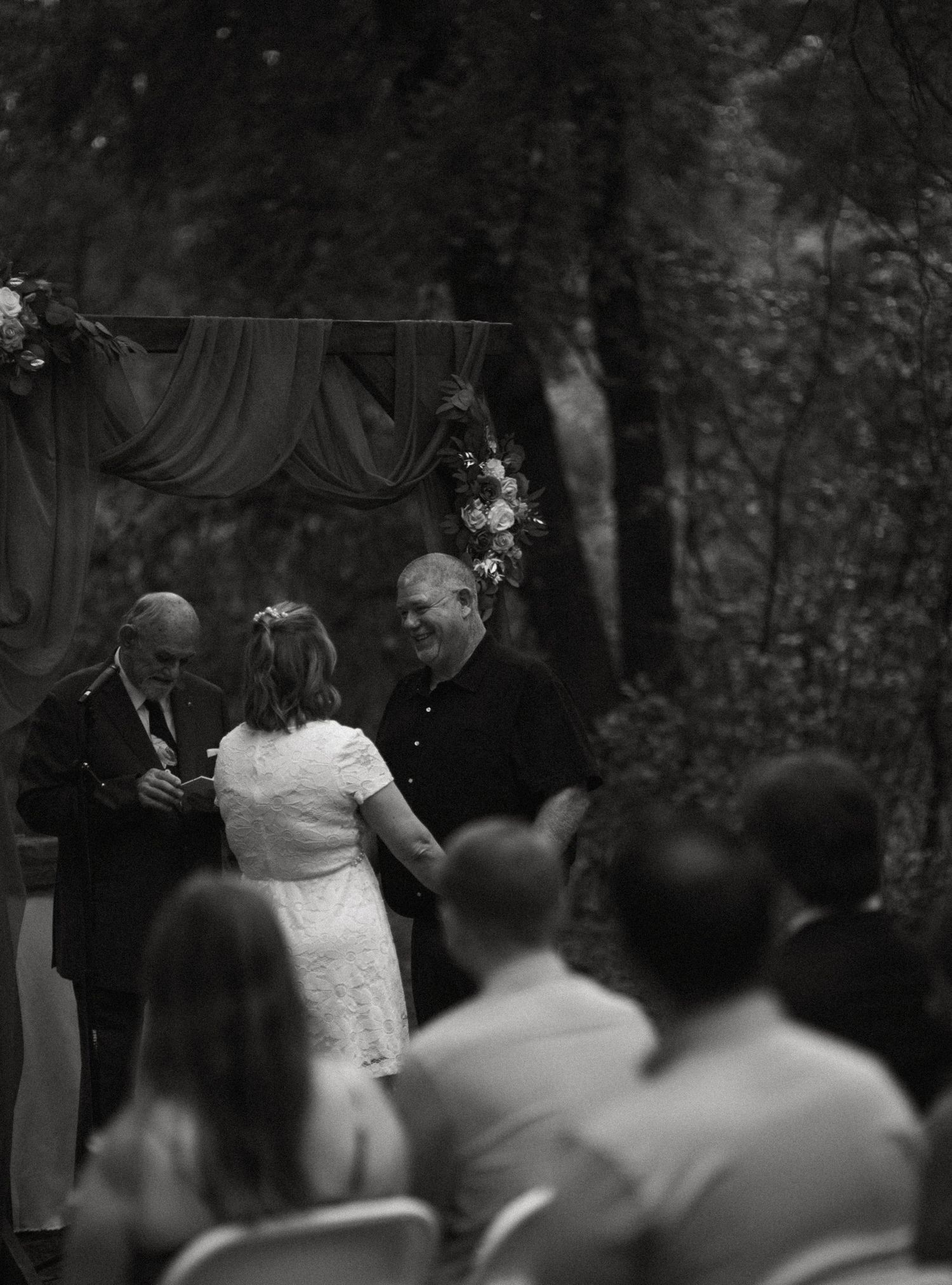Black and white photo of bride and groom exchanging vows at outdoor woodland wedding ceremony with seated guests.