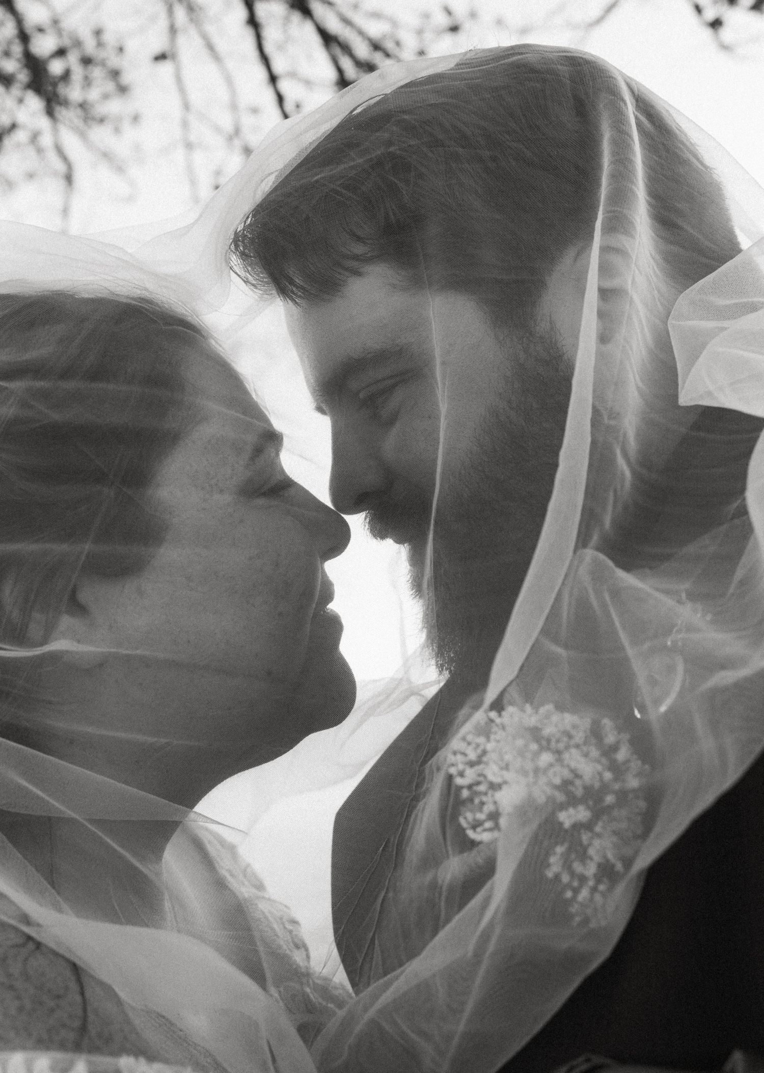 Black and white photo of bride and groom sharing a romantic moment under a flowing veil on their wedding day.