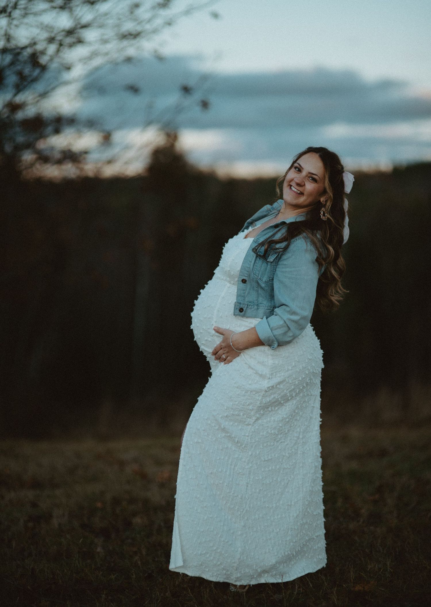 Smiling pregnant woman in white textured dress and denim jacket poses outdoors at dusk holding baby bump.