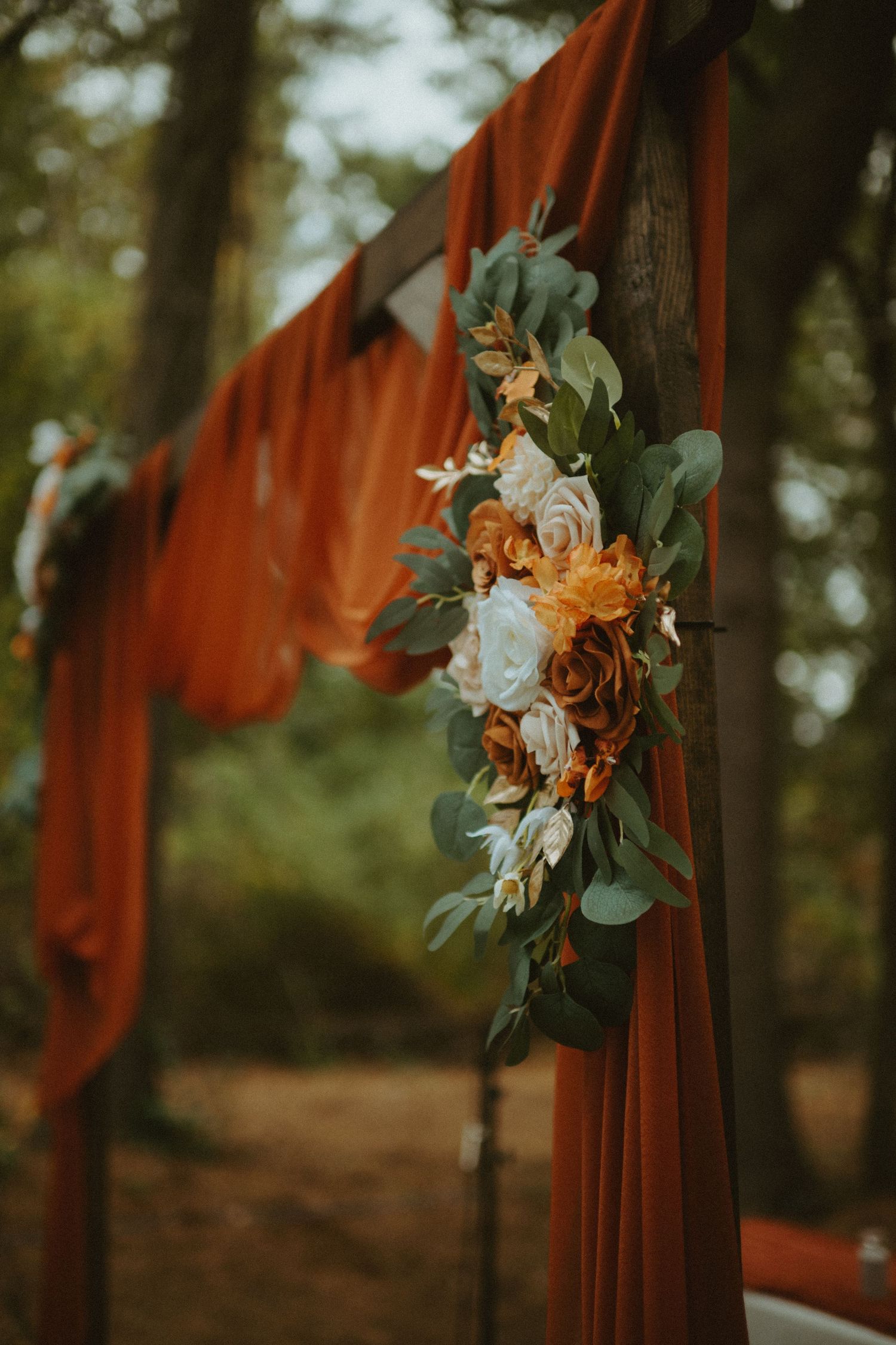 Rustic wooden wedding arch decorated with burnt orange fabric draping and autumn floral arrangement of roses and eucalyptus.
