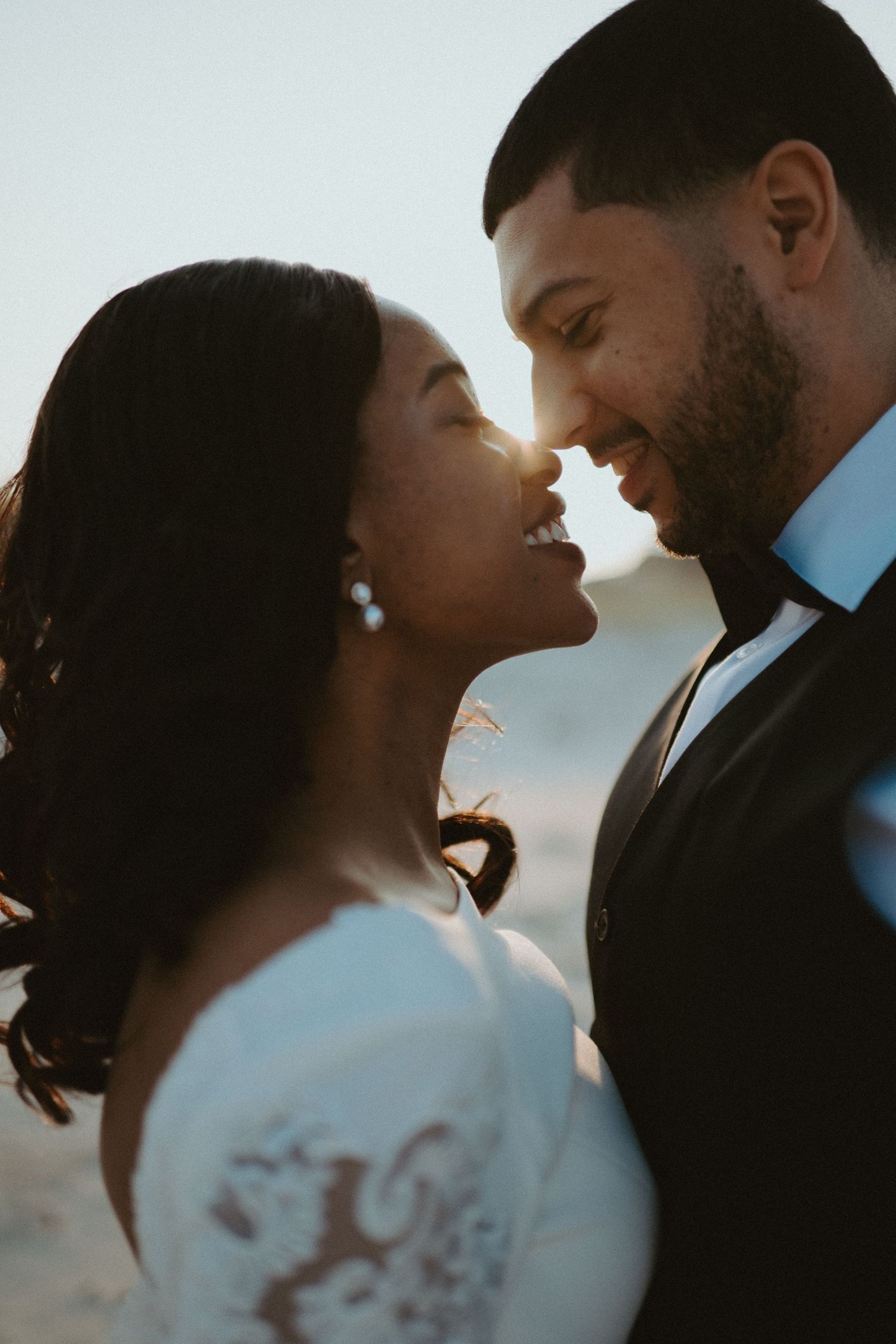 Romantic close-up of bride and groom nearly kissing at sunset, bathed in golden backlight on beach.
