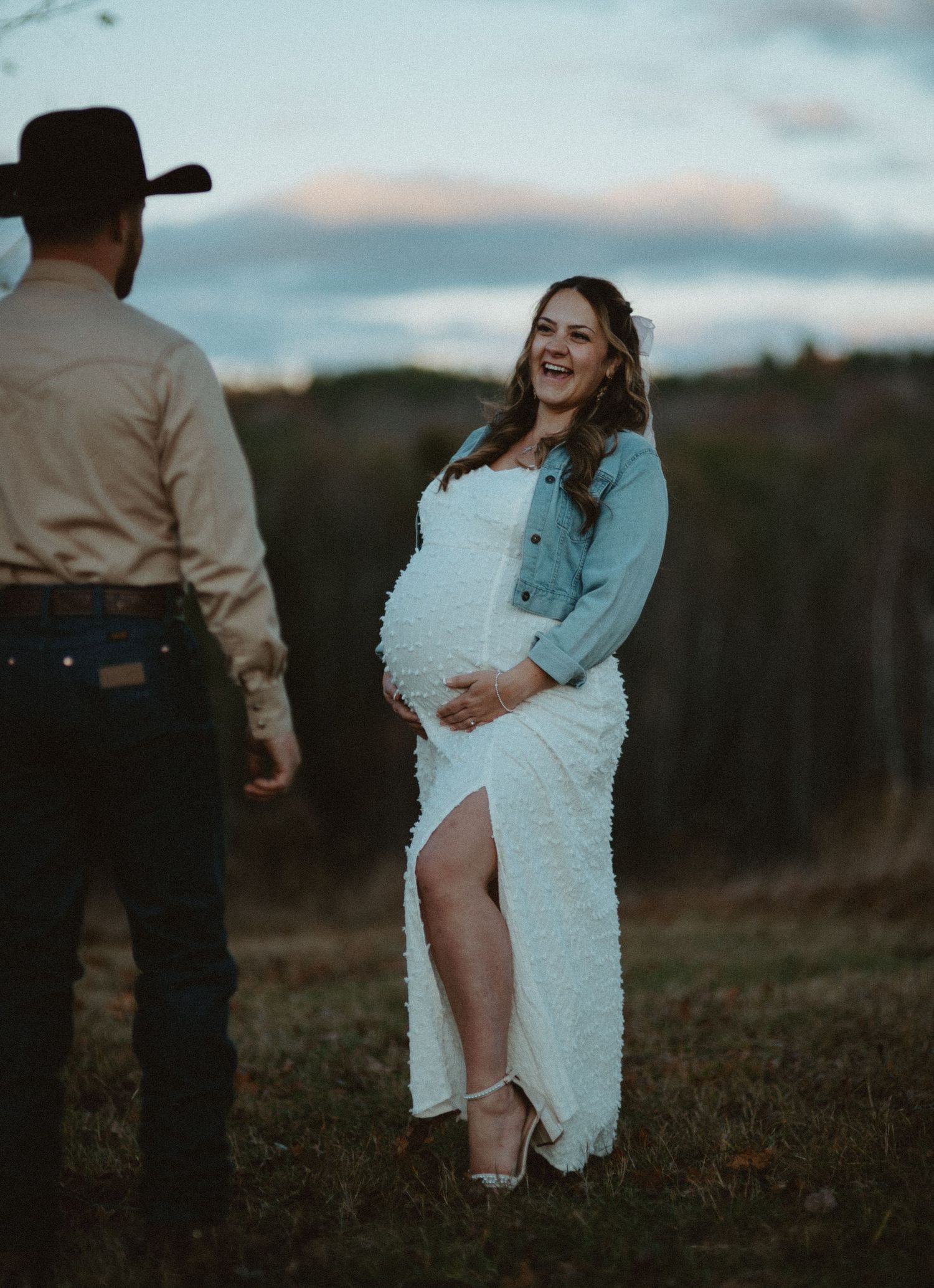 Pregnant woman in white lace dress and denim jacket laughs outdoors at sunset with husband in cowboy hat.