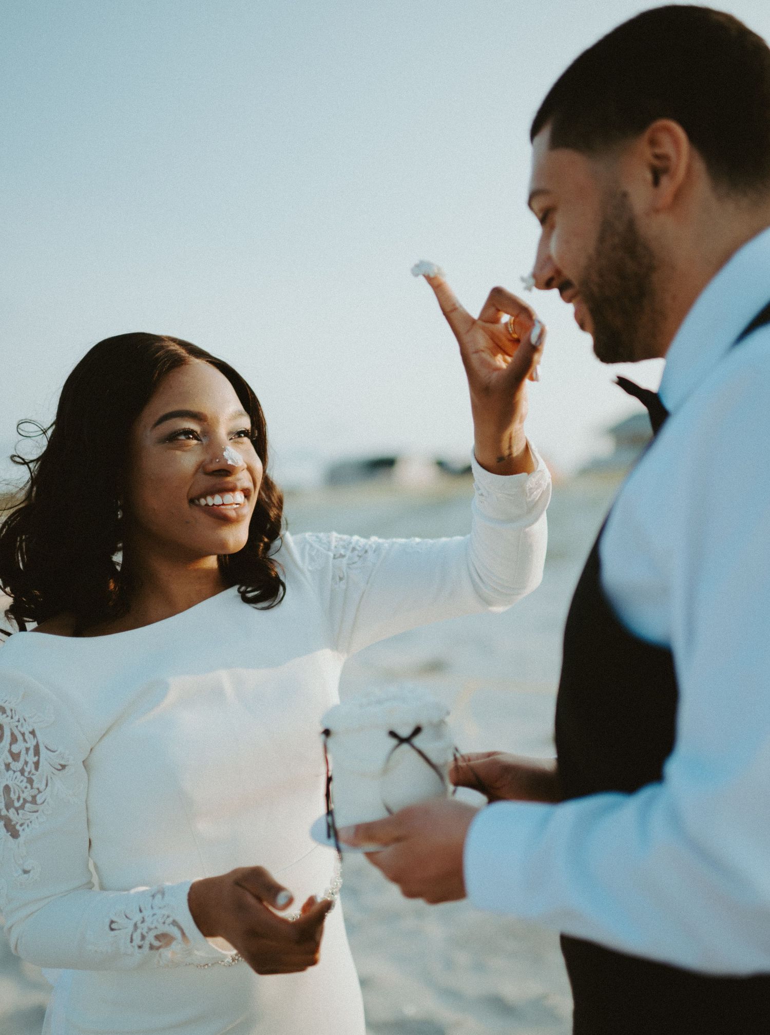 Smiling bride in white lace gown places frosting on grooms nose while on sandy beach.
