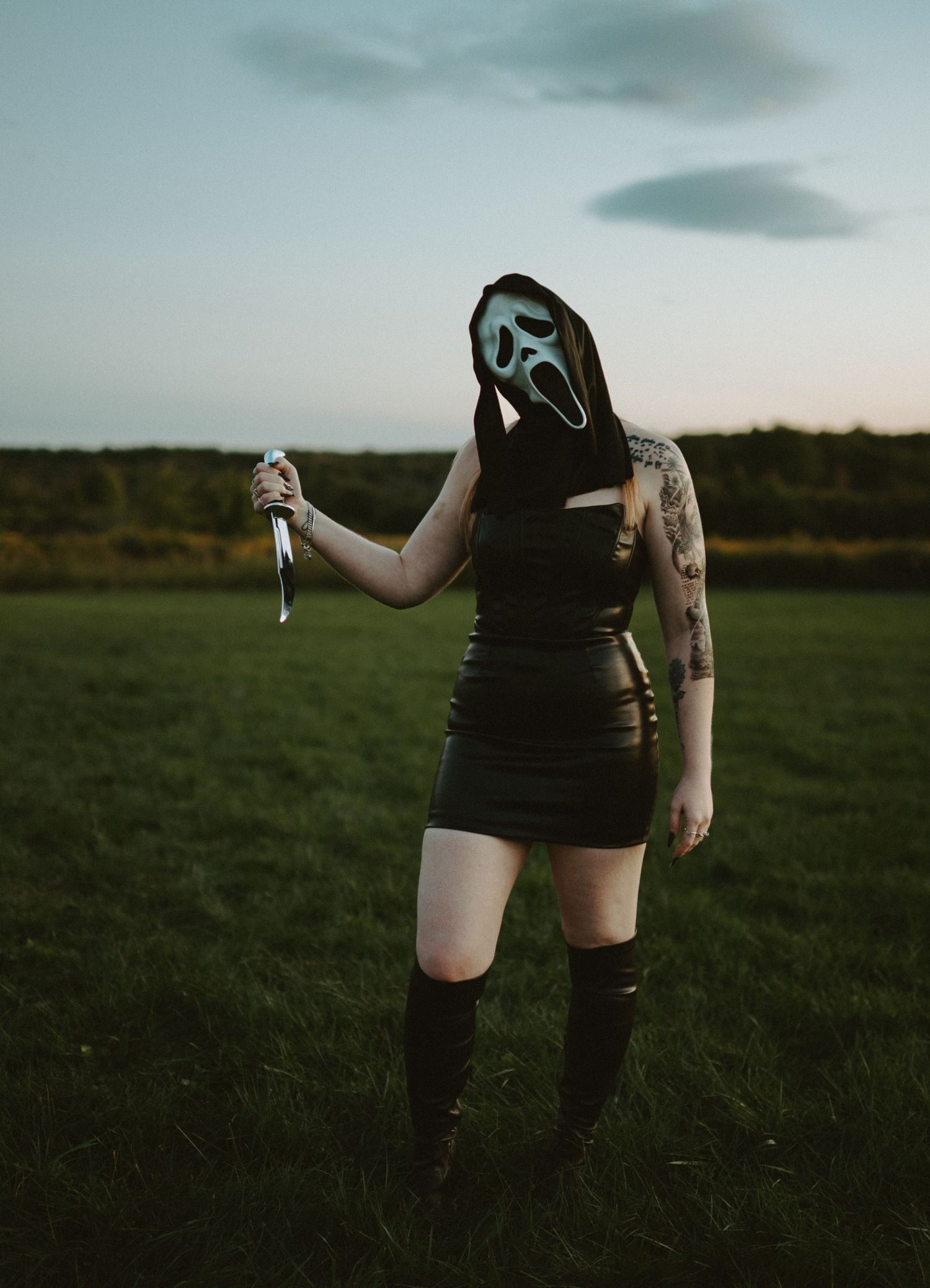 Woman in black leather skirt and Ghostface mask holding a knife in a field at dusk.