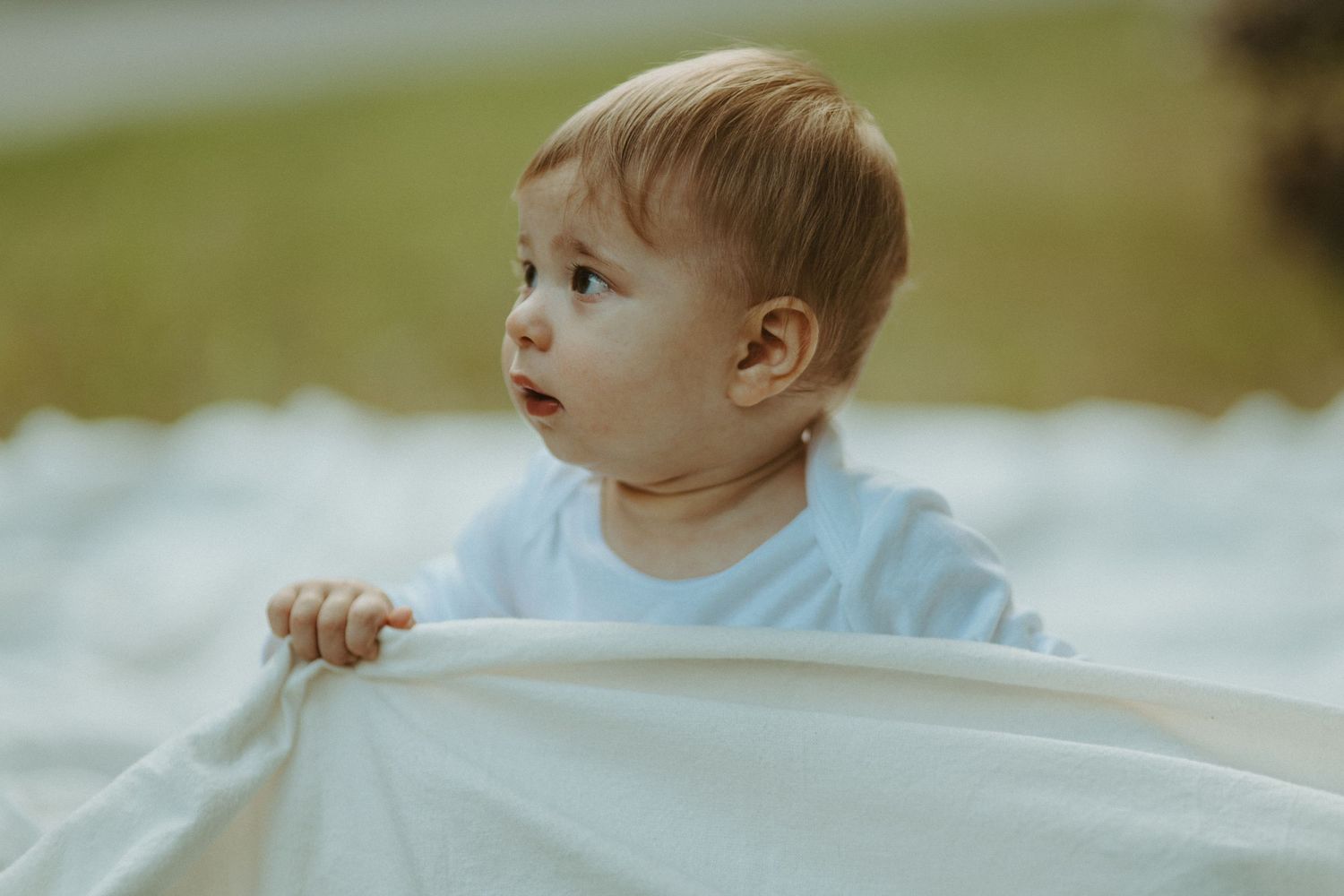Curious toddler in white shirt peeks over the edge of a white blanket outdoors.