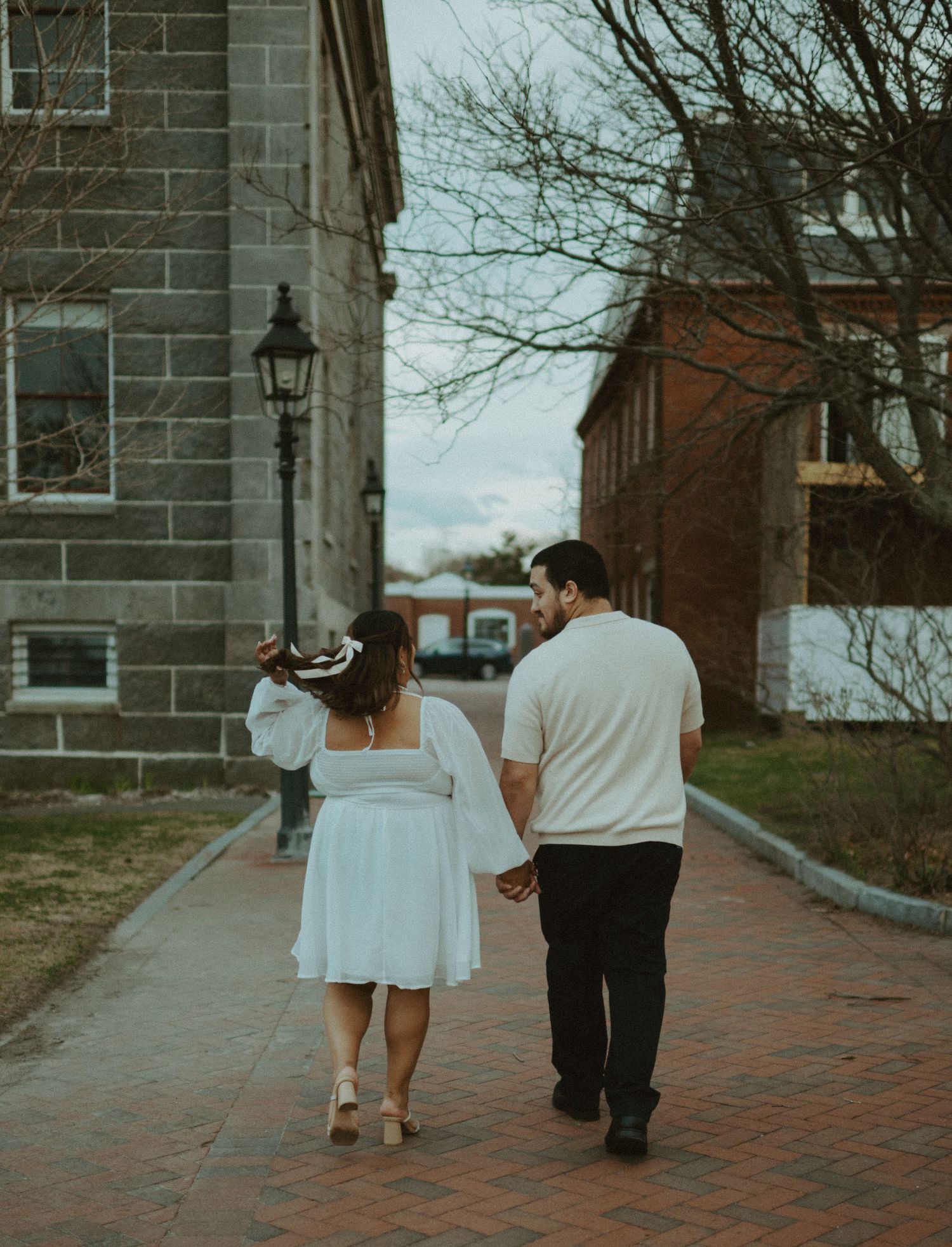Couple walking hand in hand down a brick pathway past historic stone buildings during an engagement session.