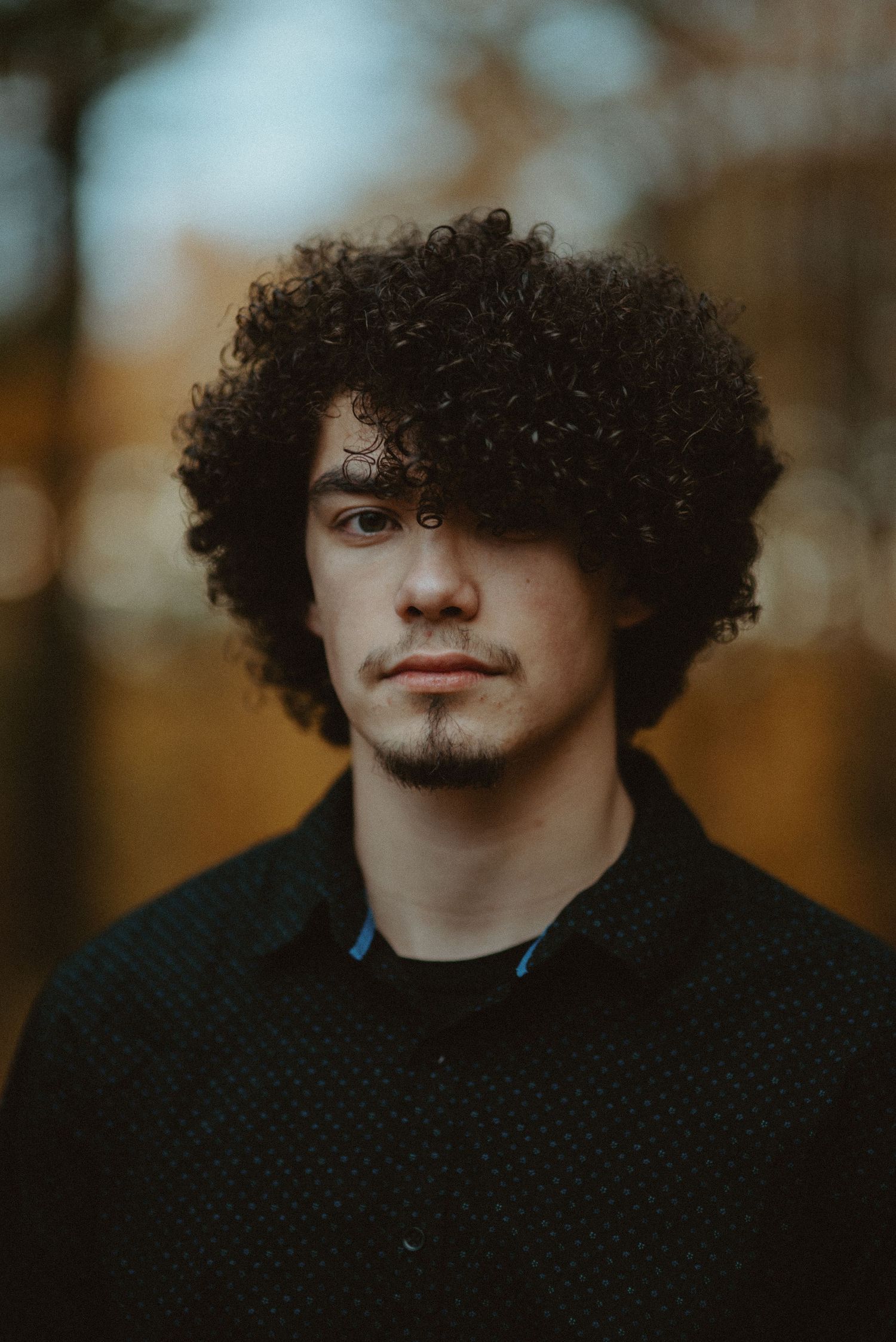 Striking portrait of young man with curly hair in dark shirt outdoors.