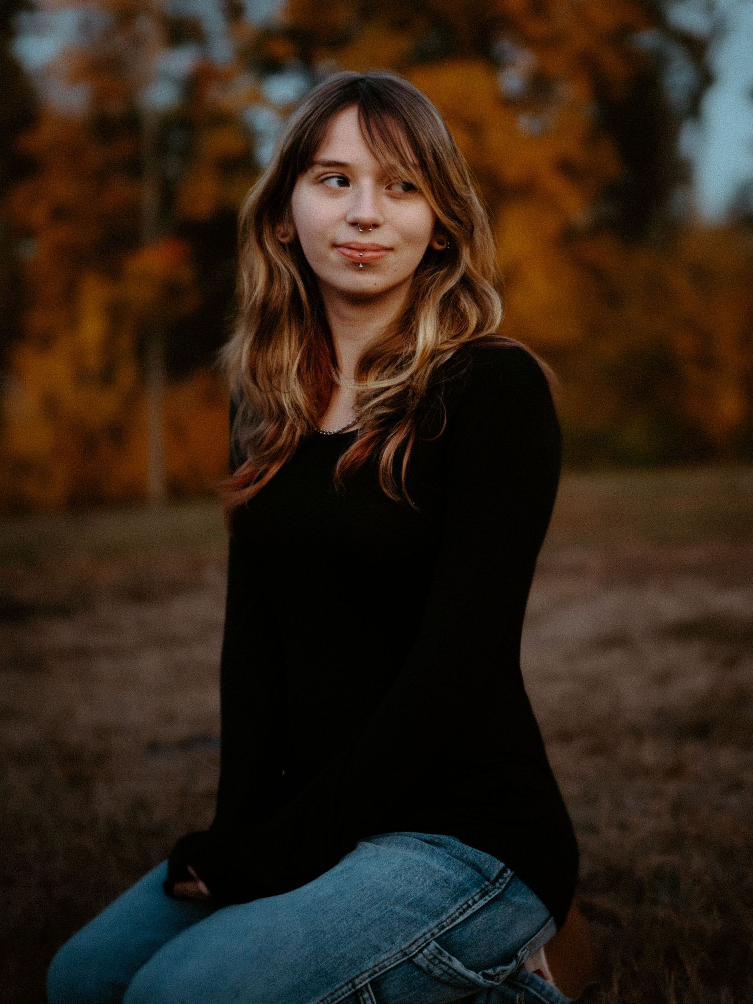 Young woman with wavy hair sitting outdoors in jeans and black long sleeve top with autumn foliage background.