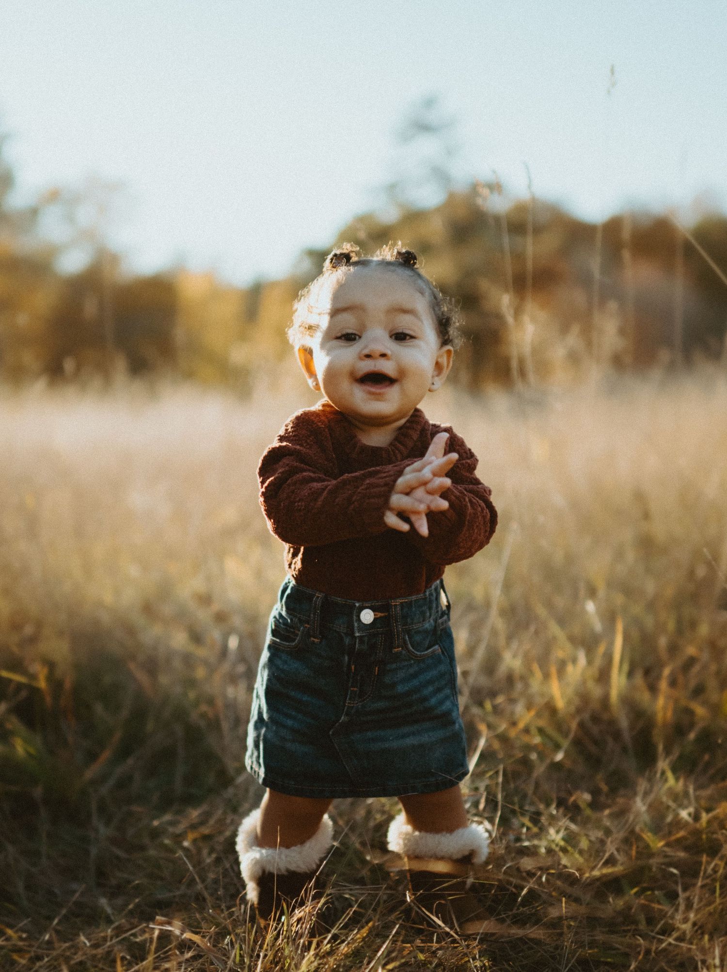 Adorable toddler girl in red sweater and denim skirt clapping hands in sunlit autumn field.