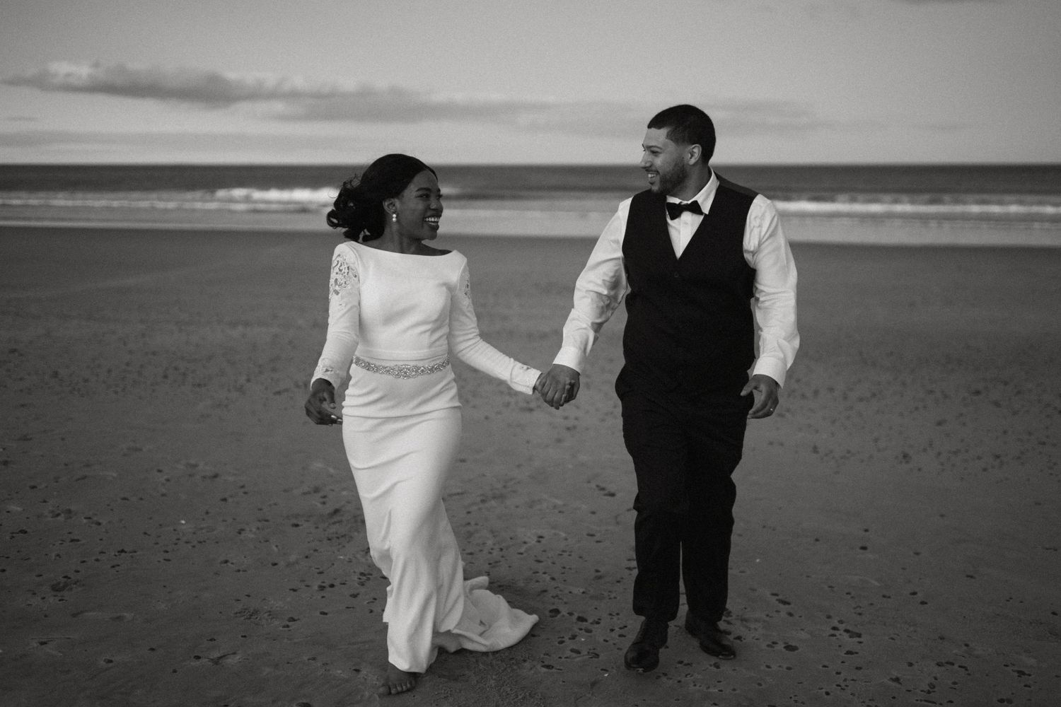 Bride and groom walk hand in hand on a beach in timeless black and white wedding portrait.