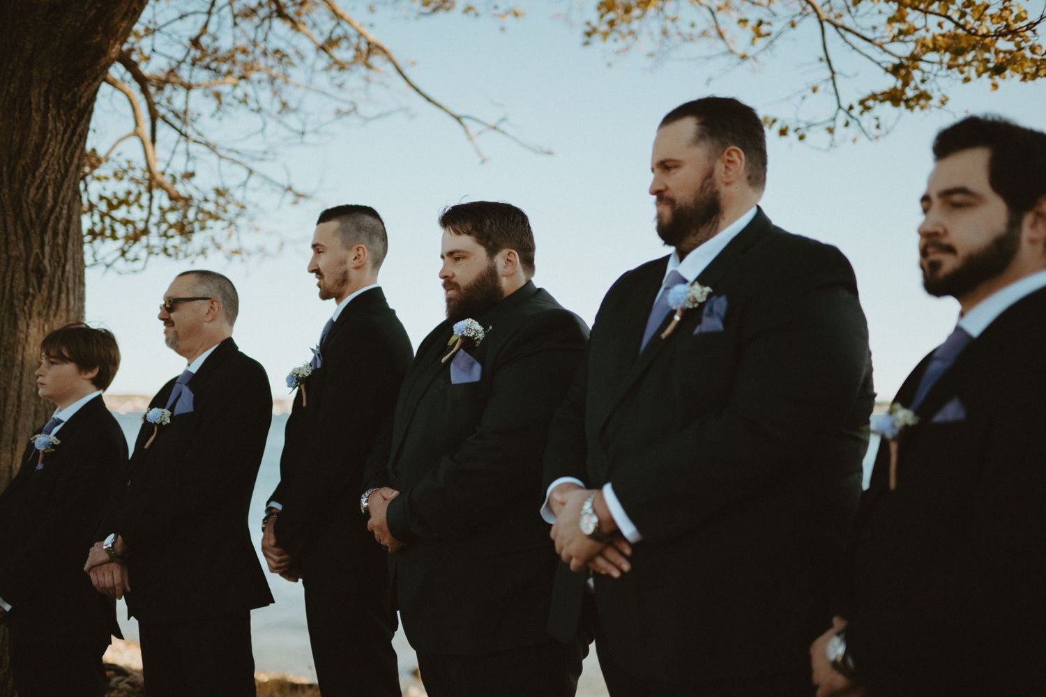 Groomsmen in black suits with boutonnieres standing outdoors under autumn trees at wedding.