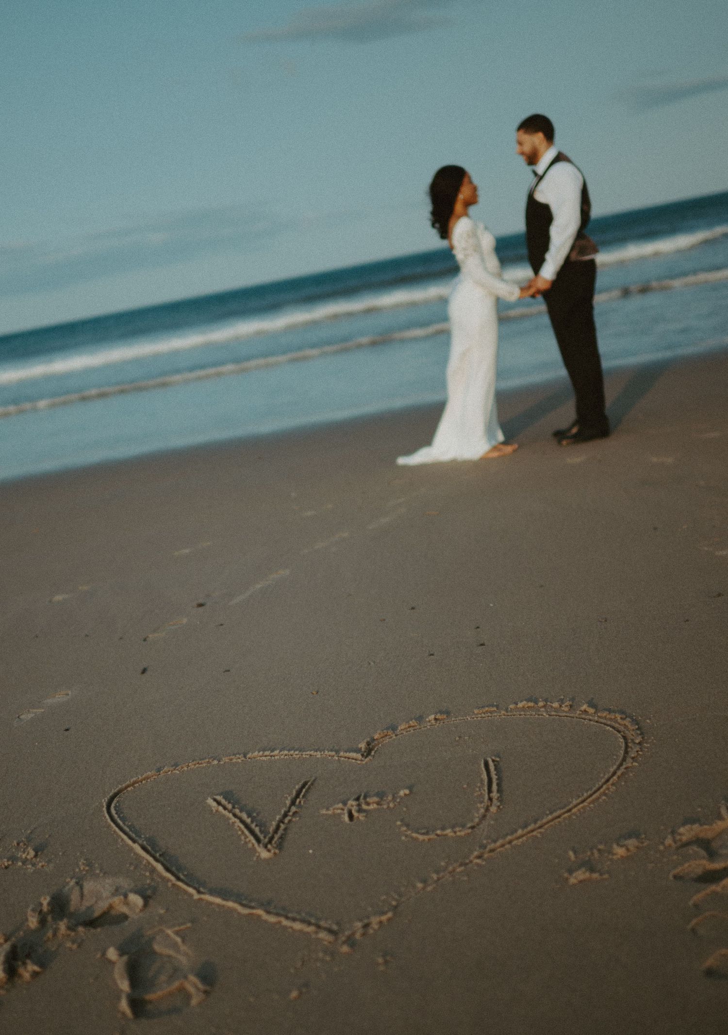 Bride and groom hold hands on beach at sunset with heart and initials drawn in sand foreground.