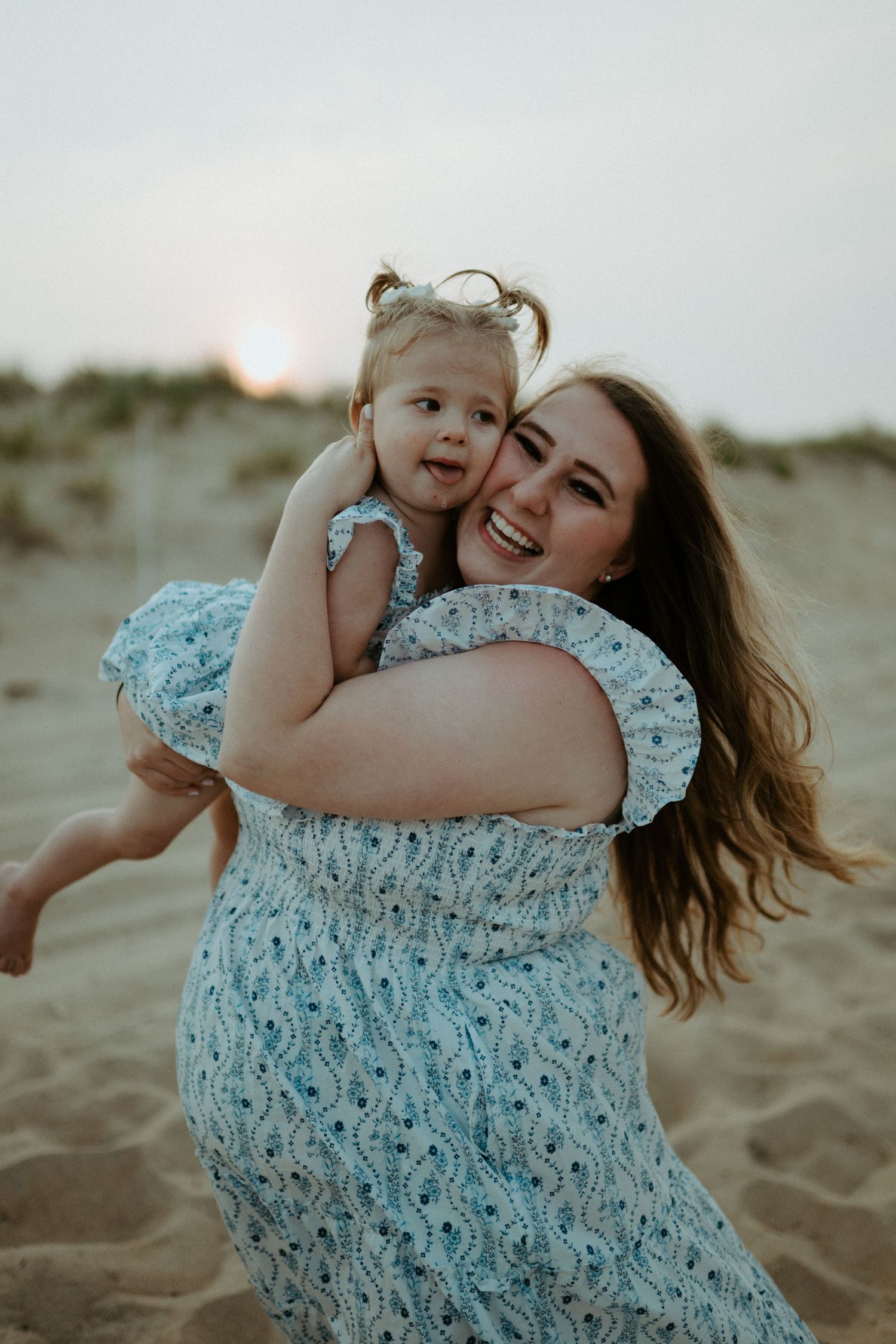 Smiling mother hugging her happy toddler daughter on a sandy beach at golden sunset hour.