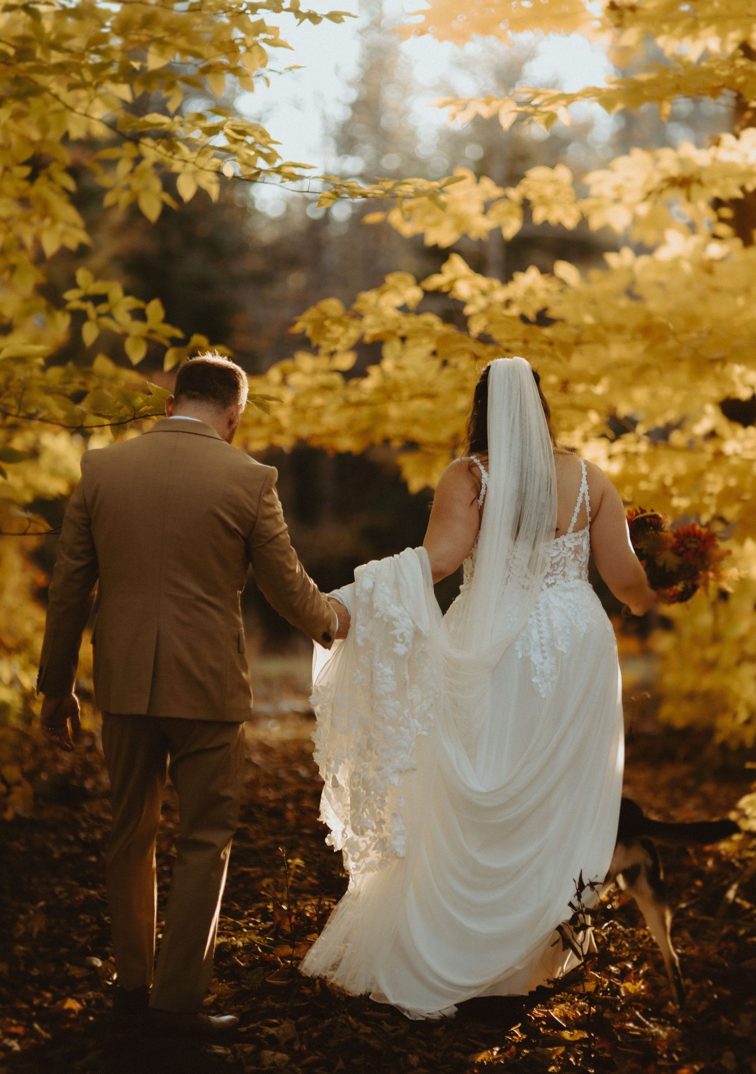 Bride and groom walking hand in hand through vibrant golden autumn foliage on their wedding day.
