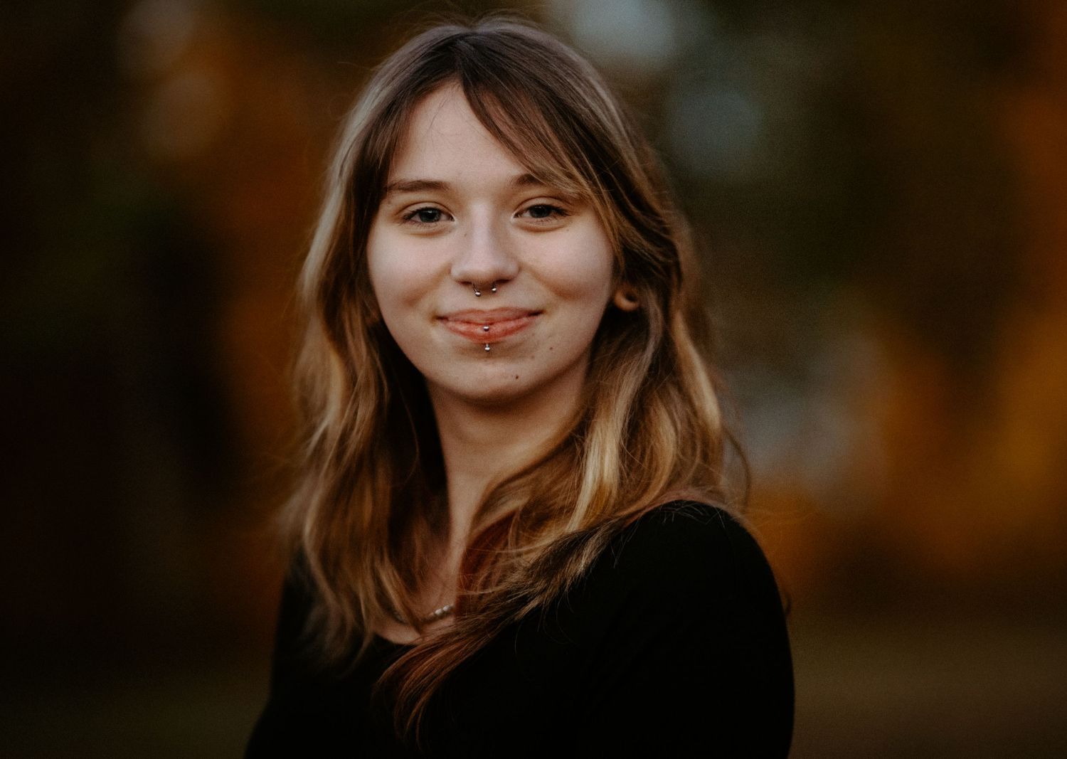 Young woman with wavy hair making a subtle smile in a black top with fall colors behind her.