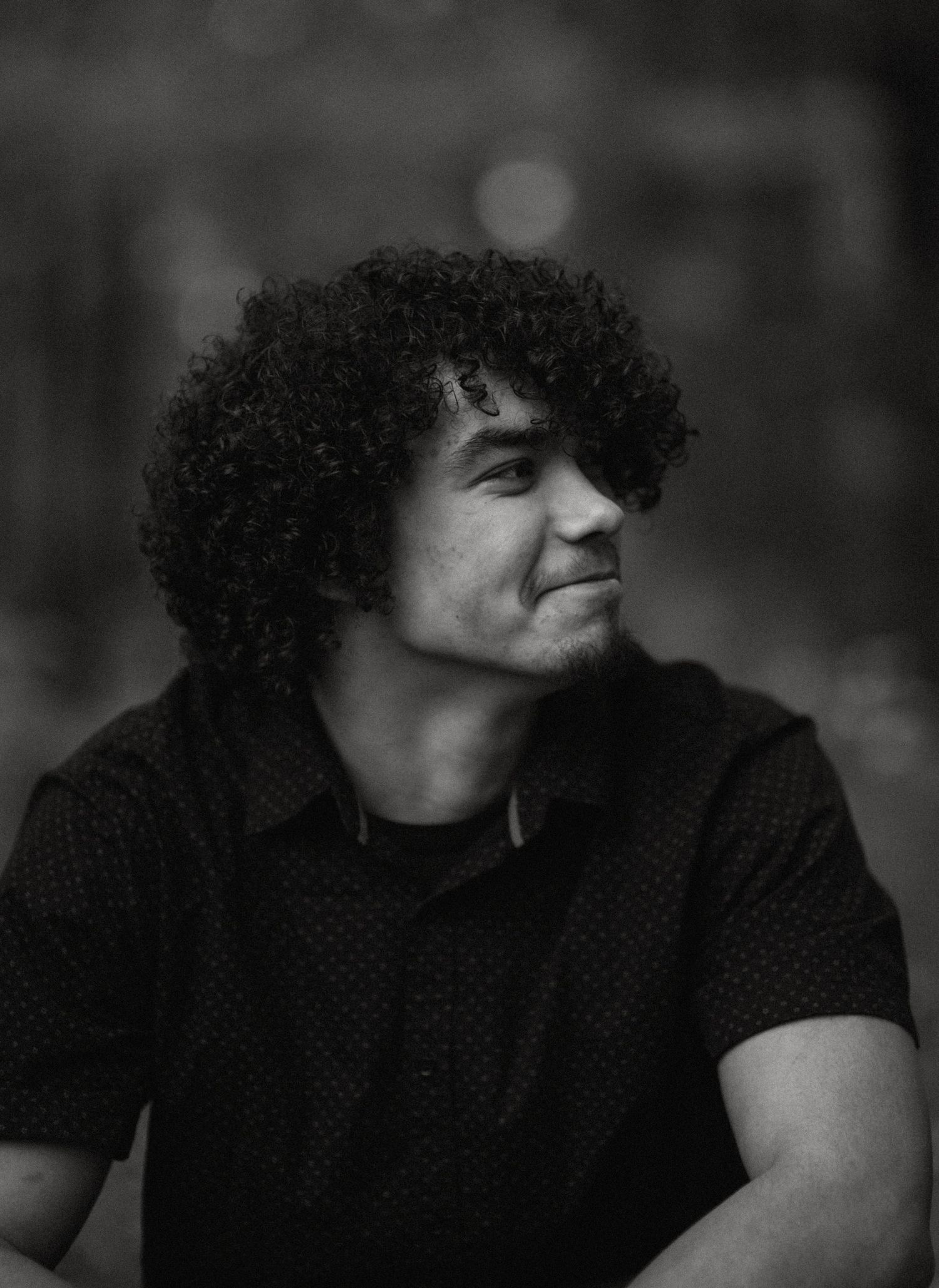 Black and white portrait of curly-haired young man smiling outdoors.
