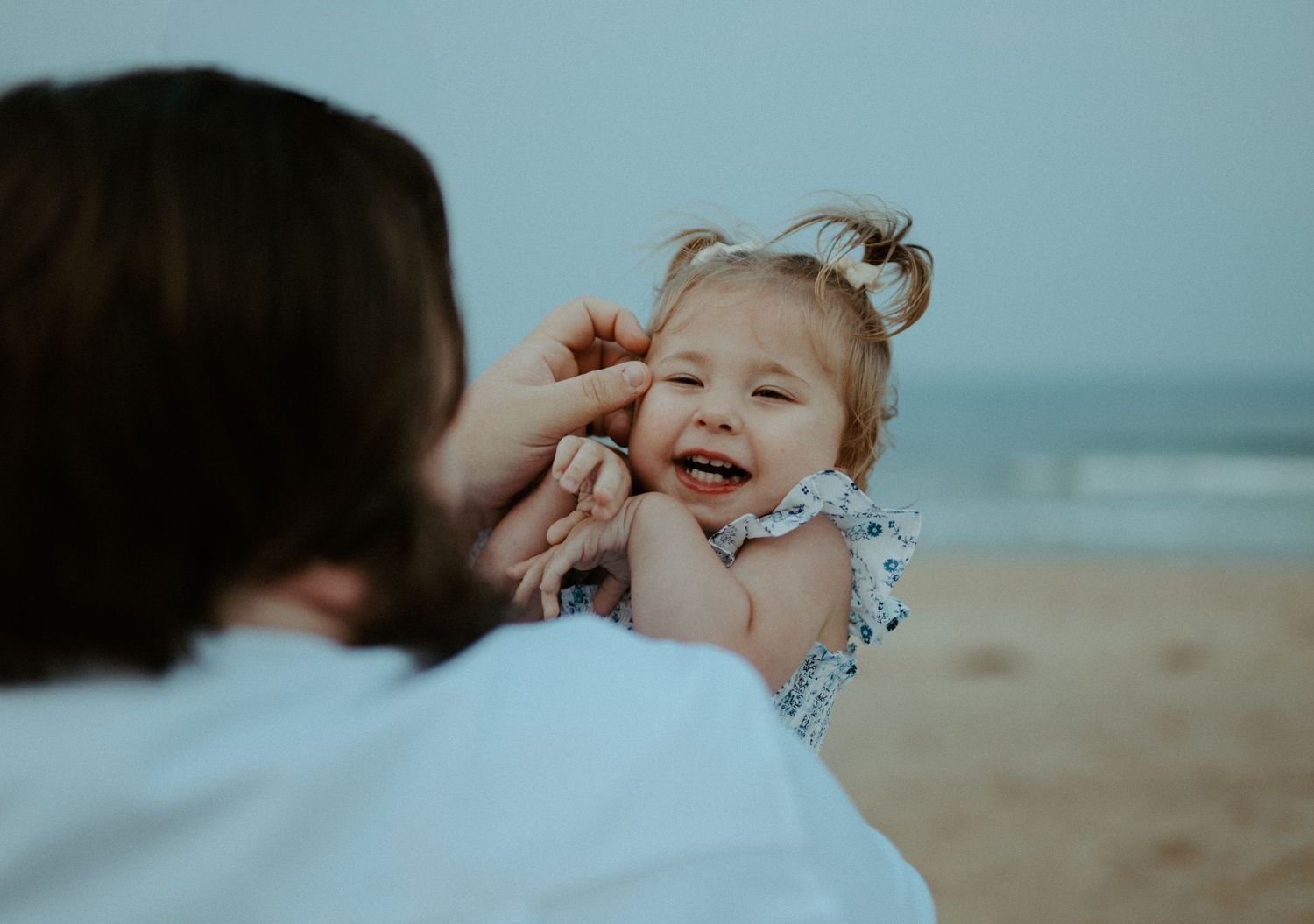 Father playfully interacts with his laughing baby girl on a sandy beach at dusk.