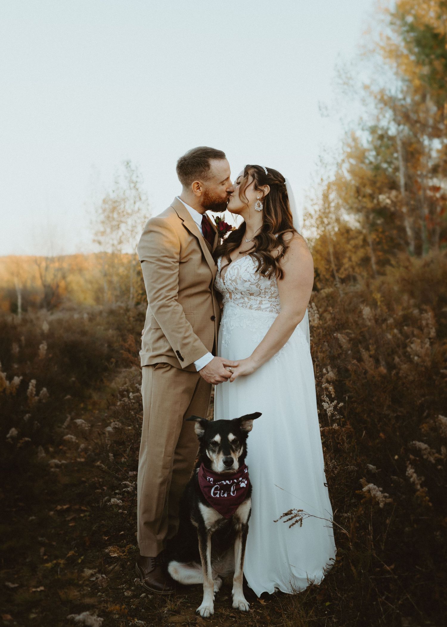 Bride and groom kissing at golden hour with their dog wearing a bandana.