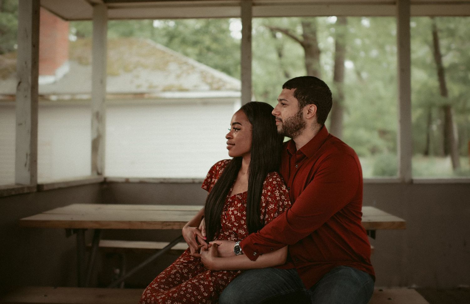 Couple sitting together on a bench in a rustic pavilion, woman in red floral dress, man in burgundy sweater.