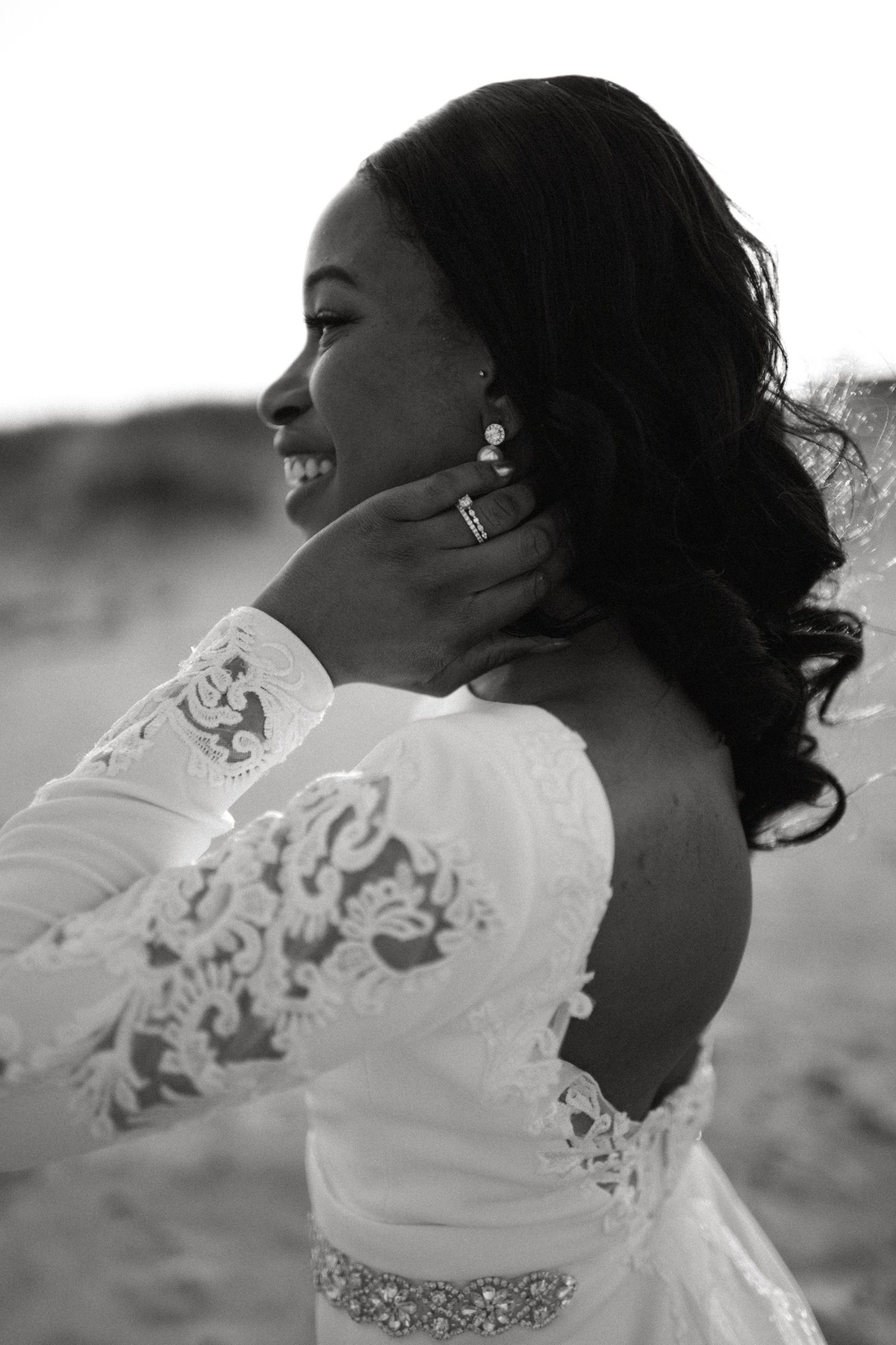 Elegant black and white portrait of a smiling bride in lace gown touching her earring outdoors.