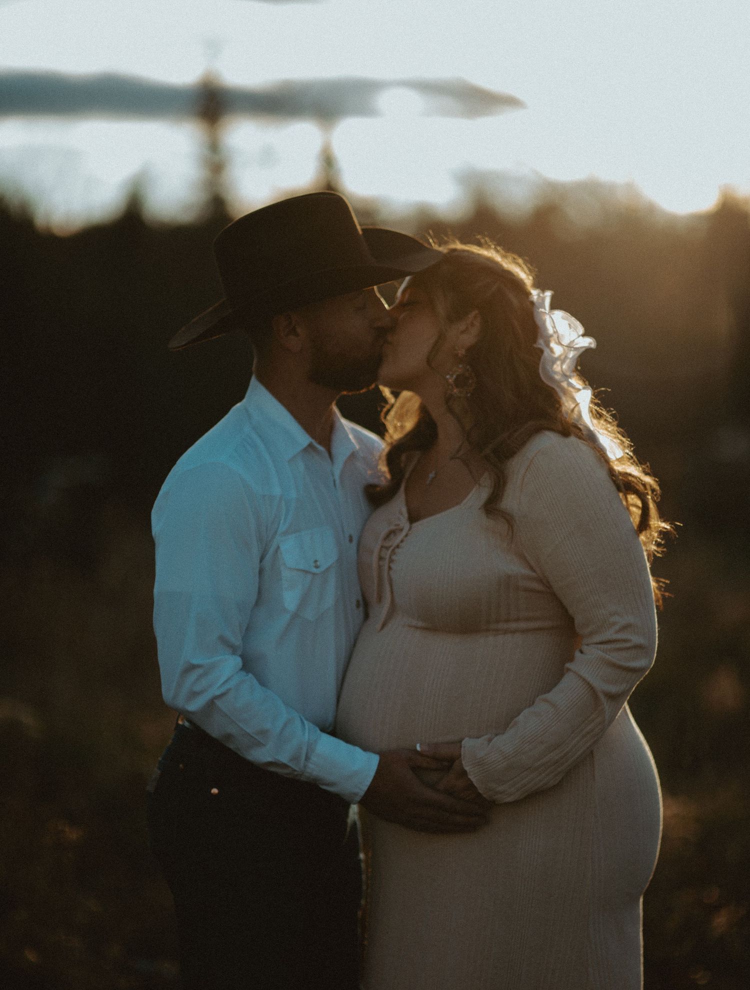 Pregnant couple shares tender kiss at golden sunset, man in cowboy hat embracing woman in white.
