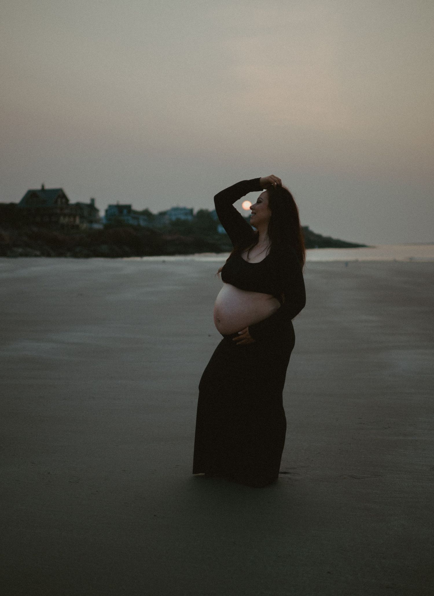 Pregnant woman in black dress poses on beach sunrise, hand in hair.