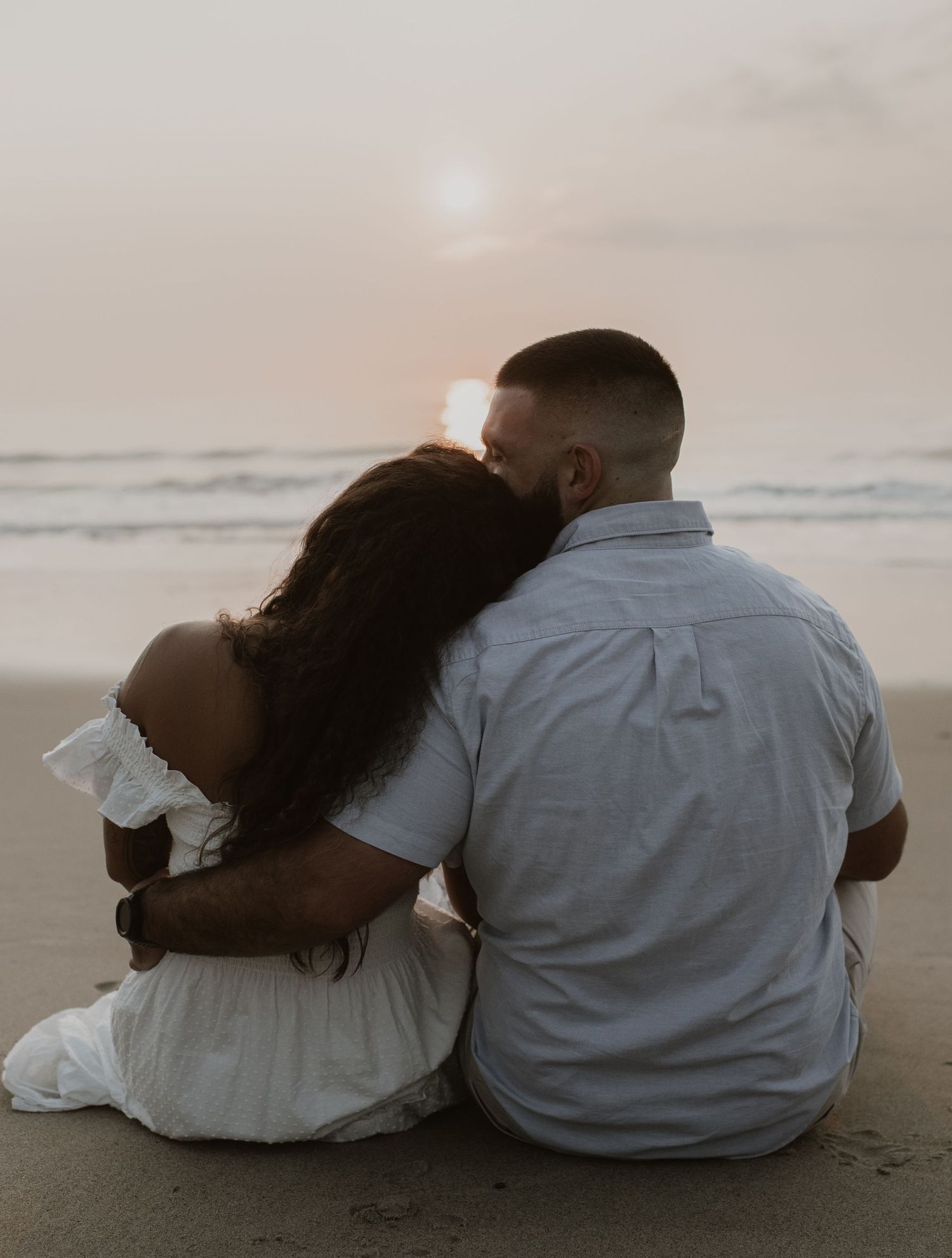Woman resting head on mans shoulder while sitting on beach watching a glowing sunset together.