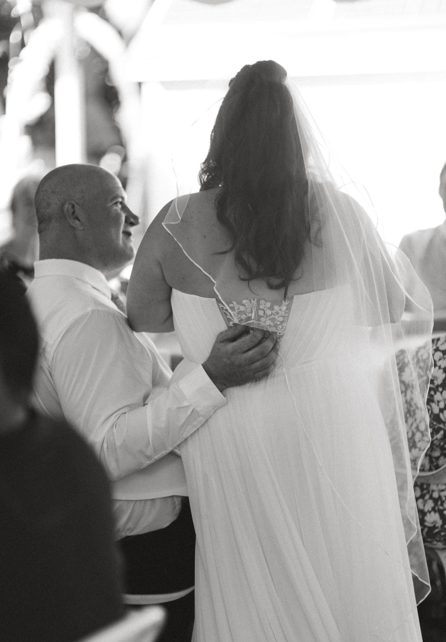 Black and white photo of bald groom embracing bride in veil during outdoor wedding ceremony.