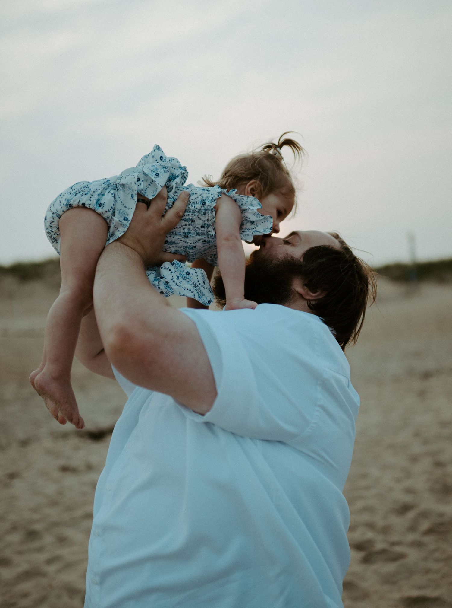 Bearded father lifts his smiling toddler daughter high in the air on a sandy beach.