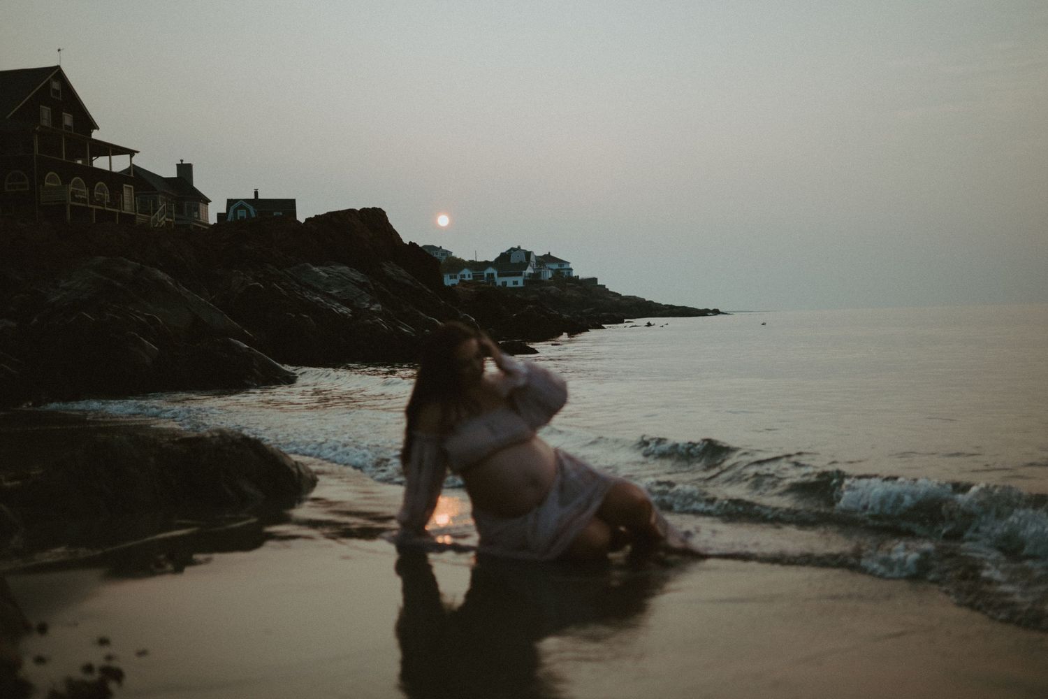 Pregnant woman in pink dress sits by ocean waves at dawn with sunrise over rocky New England coastline.