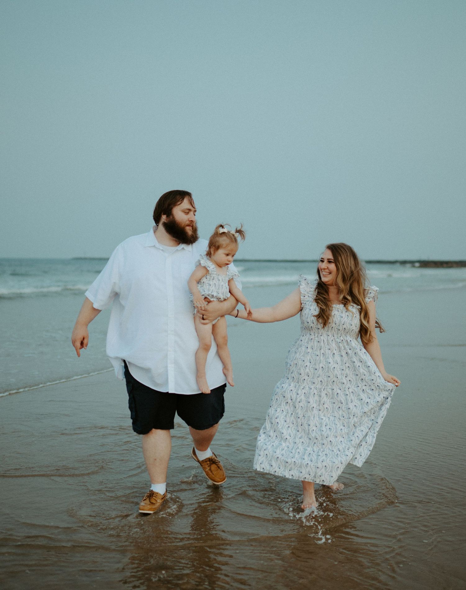 Happy family of three walking along shallow ocean waves on the beach at sunset together.