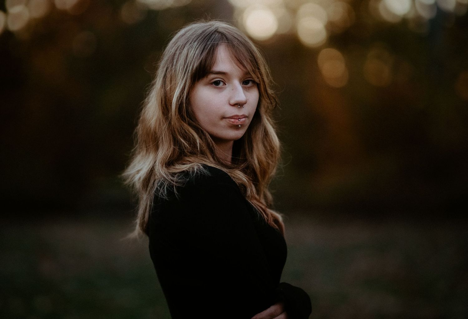Young woman with wavy hair posing with arms crossed in a black top against a soft golden hour background.