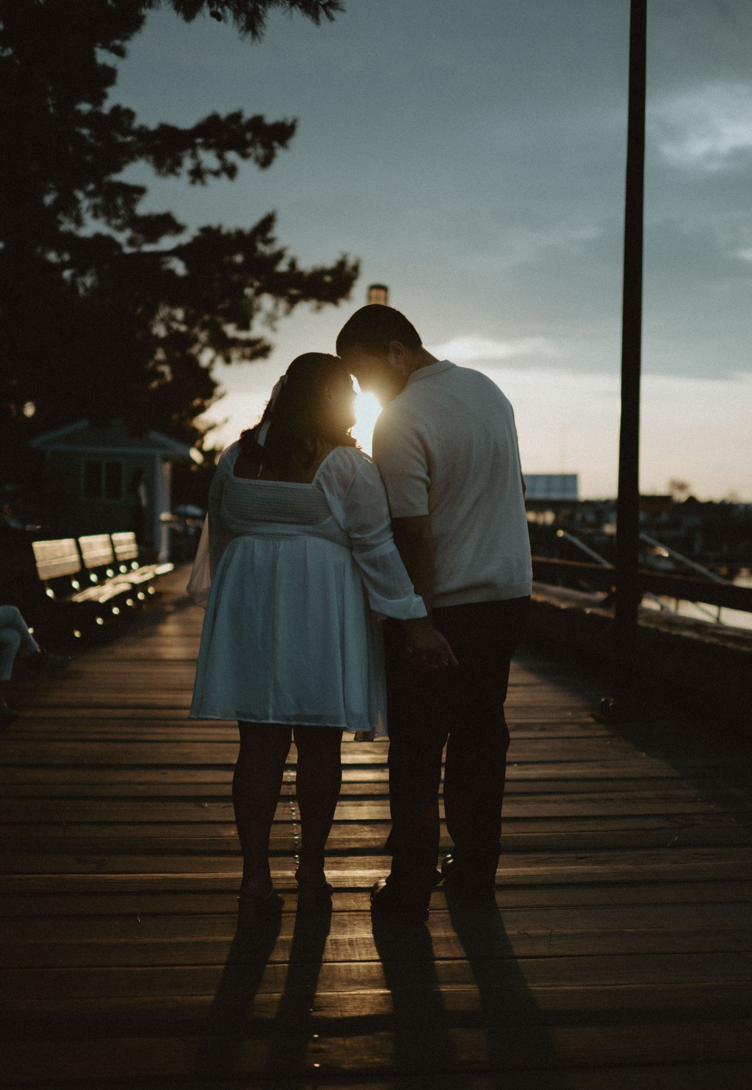 Romantic couple silhouetted against a glowing sunset on a wooden boardwalk by the waterfront at dusk.